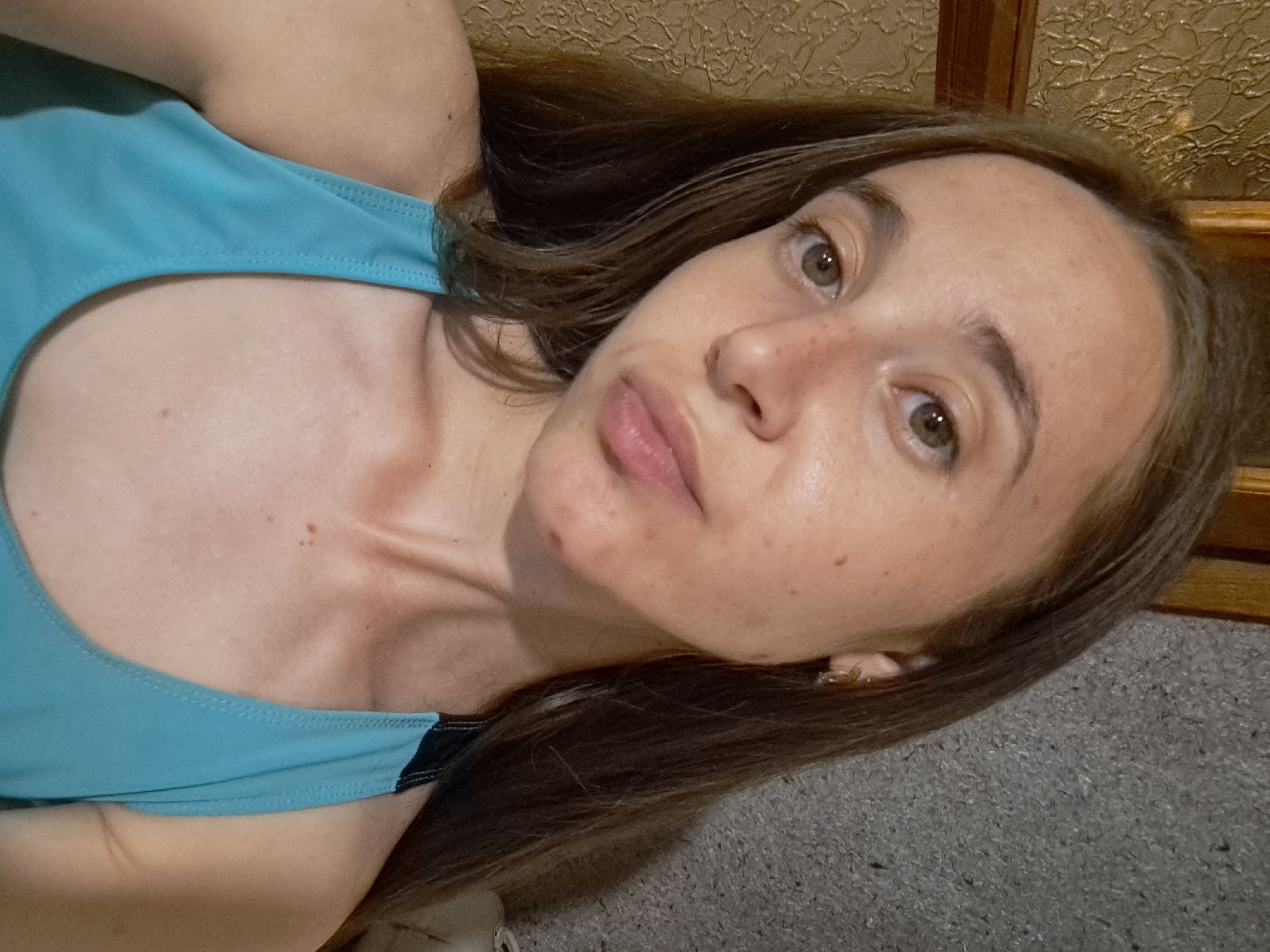 A young woman with long brown hair is taking a selfie in a room with textured walls and wooden trim. She is wearing a bright blue tank top. Her expression is neutral. The background appears to be an indoor residential setting.