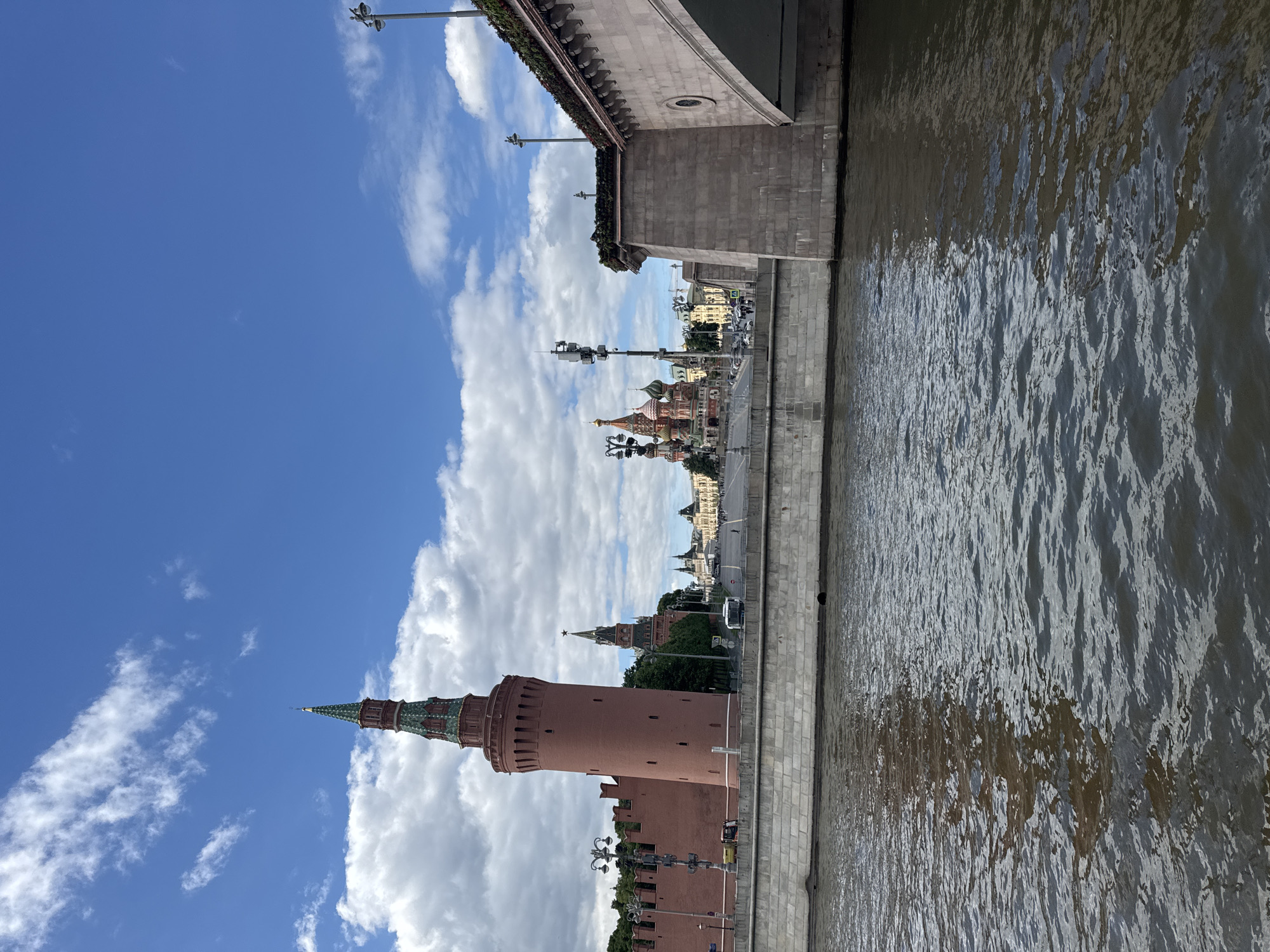 The image presents a view of Moscow, Russia, showcasing a river in the foreground with shimmering water. Adjacent to the river is a stone embankment. On the other side of the embankment, there are various structures, including a prominent red brick tower from the Moscow Kremlin, part of the historic fortified complex. Beyond this, there are buildings, including St. Basil's Cathedral and other structures lining the landscape. The sky is blue with scattered white clouds. A bridge is partially visible in the upper part of the image, and lampposts are visible on both the river side and the Kremlin side.