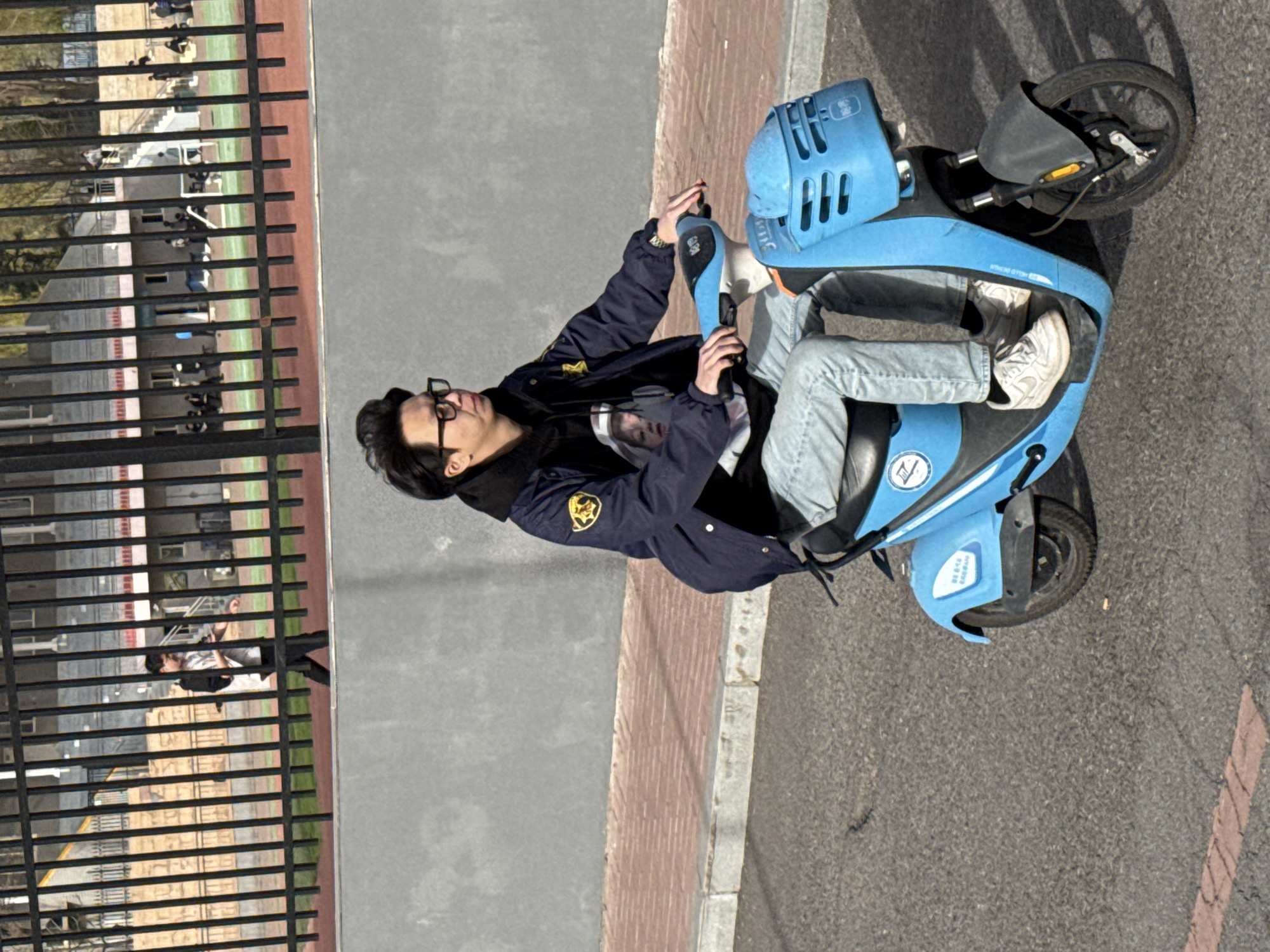A person wearing eyeglasses, a dark jacket with a yellow badge, light-colored jeans, and white sneakers is seated on a light blue scooter on a paved surface with cracks and a painted line. The scooter has black tires and appears to be a rental. A sidewalk with red brick borders the paved surface. To the left is a grey concrete wall next to a tall, black, metal fence. A grassy area and a building are visible behind the fence. This scene is in Zhongtun, China.
