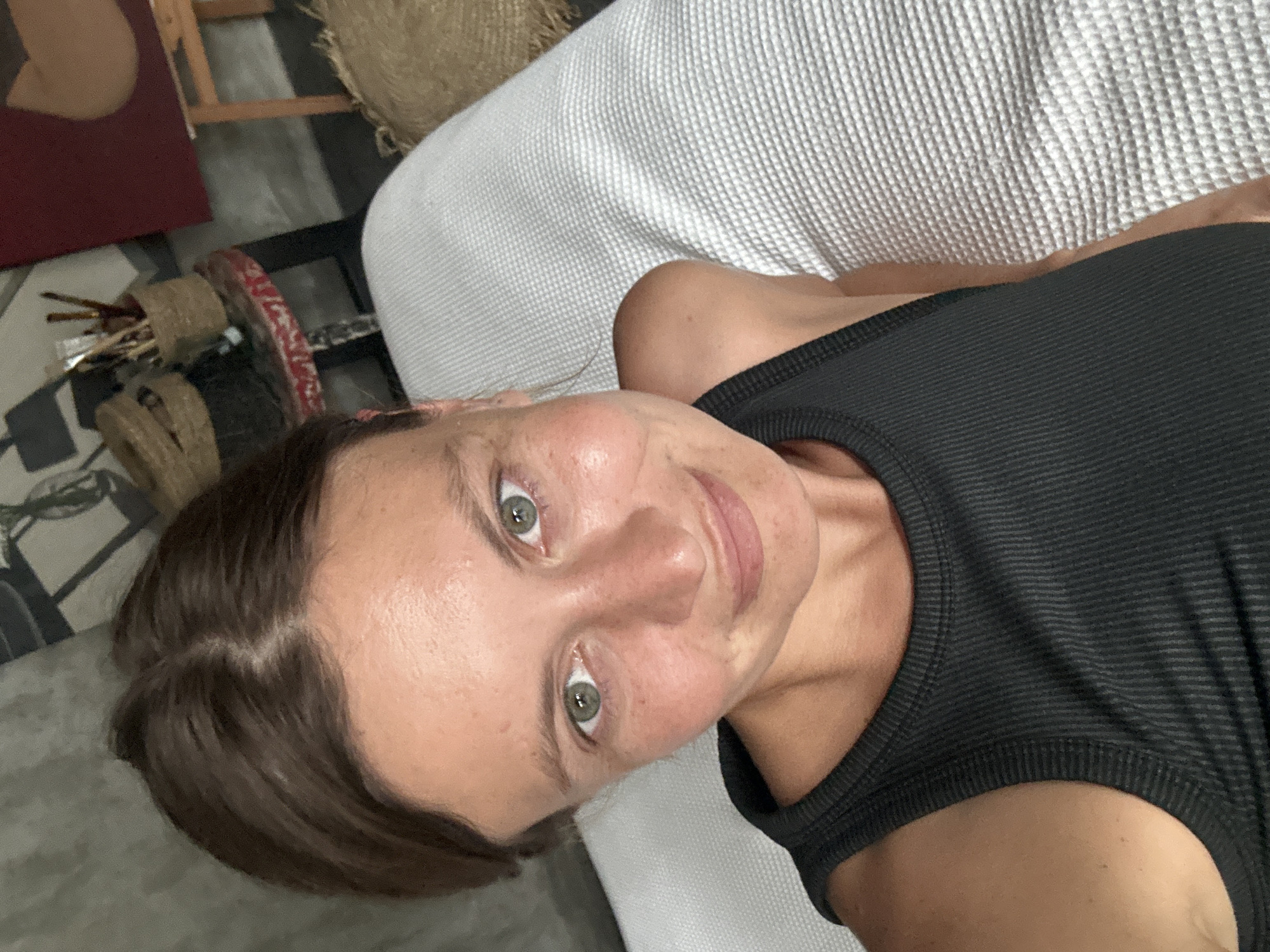 A young adult woman is captured in a close-up, high-angle shot, looking directly at the camera with a gentle, engaging smile. Her light green eyes are prominent, and her brown hair is parted in the middle. She has fair skin with subtle freckles or blemishes. She is wearing a dark, ribbed tank top, suggesting a casual or athletic attire. The image is rotated 90 degrees clockwise, making her appear to be lying on a light-colored, textured surface, possibly a sofa or bed.
The background suggests an indoor setting, likely a room with artistic or craft elements. To her left, behind her head, there is a wooden easel or frame, a reddish-brown canvas or board, and some woven baskets. Further in the background, a dark floor is visible, and on it, a container filled with art brushes or similar tools sits near a red-rimmed object. The overall lighting is soft and natural, indicating daytime indoors. There is no visible text in the image. The scene conveys a relaxed and casual atmosphere, possibly in North Kuta, Indonesia.