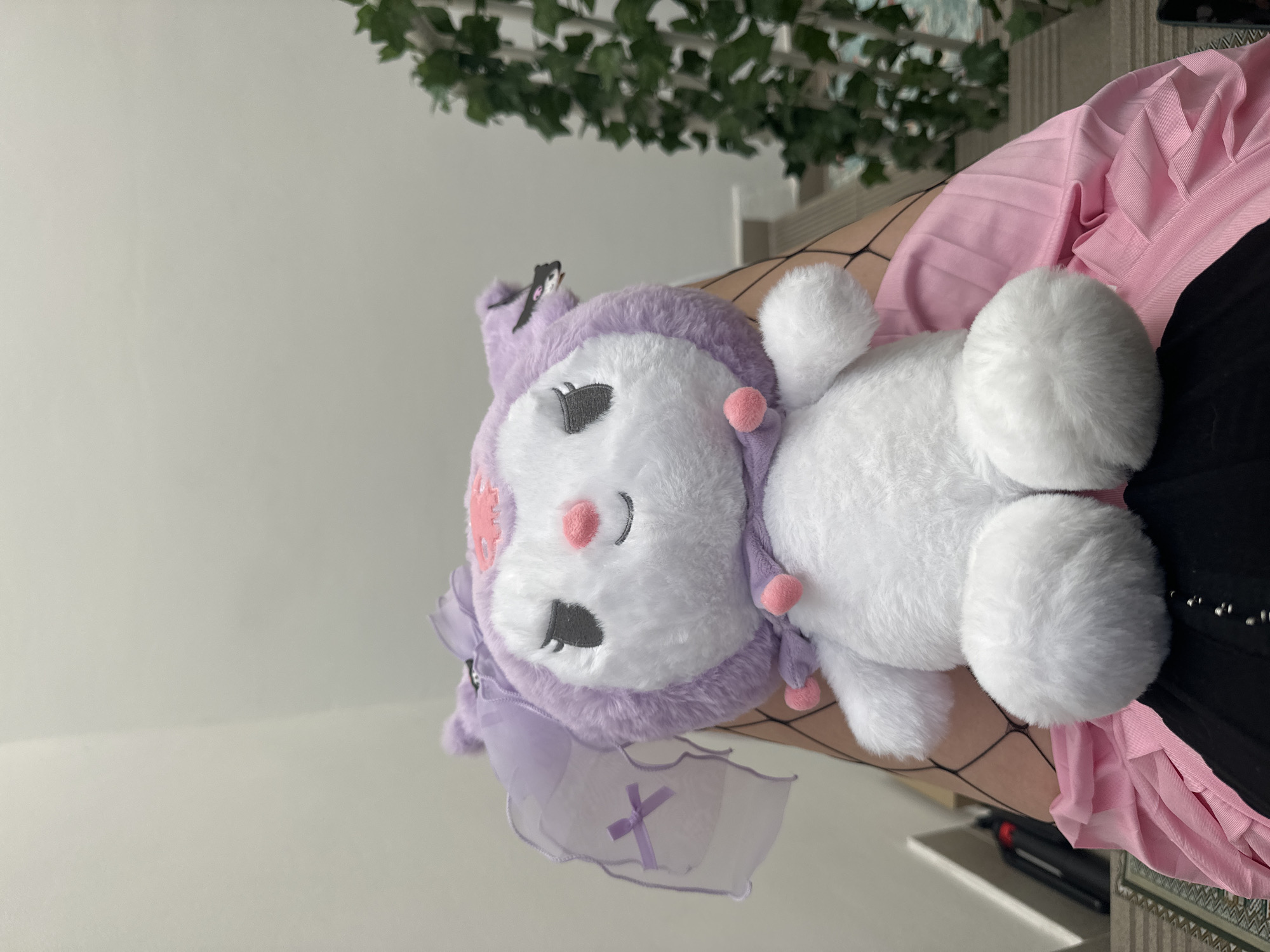 A plush toy that resembles a white rabbit with a purple and white head and a nurse cap lies on the lap of a person who is wearing fishnet stockings, a pink pleated skirt, and a black top. The scene is indoors, with a white wall visible in the background, and an artificial green ivy garland hanging near the top. The location appears to be City of Zagreb, Croatia.