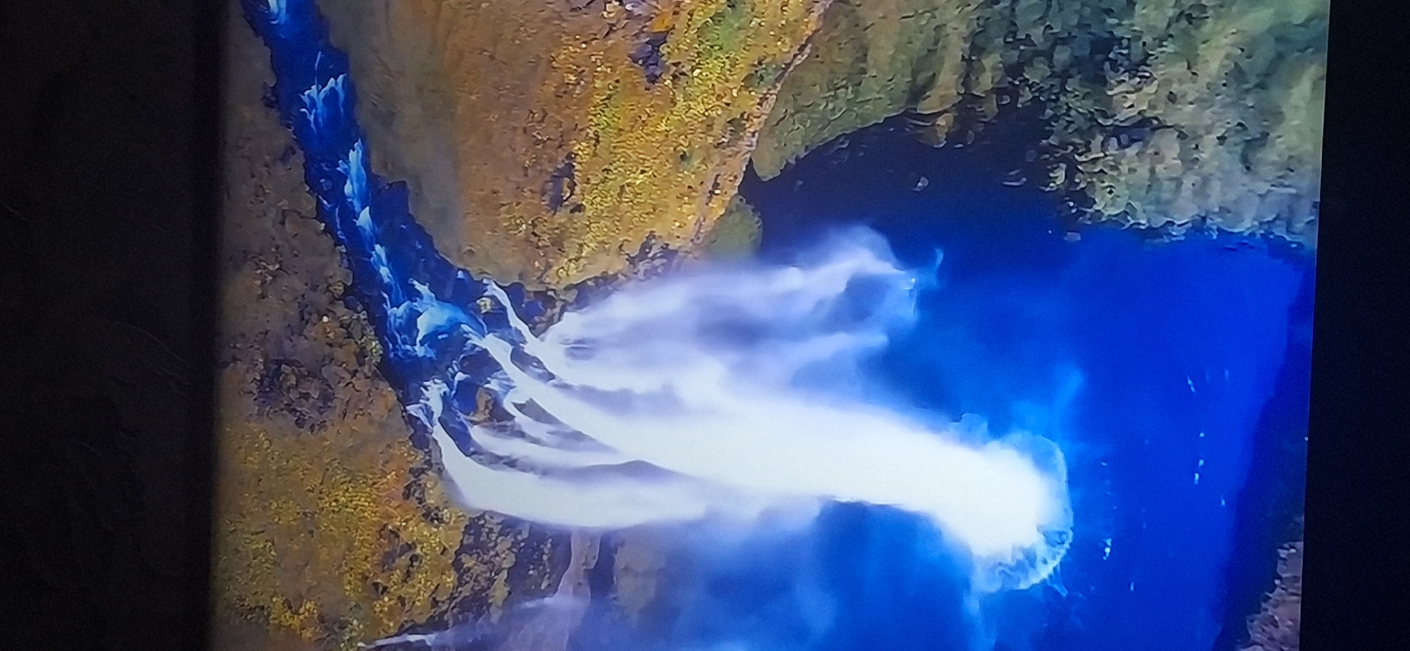 This is an aerial, slightly tilted, close-up shot of a large waterfall. The water is white and frothy, cascading down from a rocky cliff face covered in yellowish-green vegetation. The water plunges into a deep blue pool below, where it churns and creates a white mist. A river of bright blue water flows away from the waterfall, disappearing into the distance. The scene is rich in natural elements, with the vibrant colors of the water and vegetation creating a striking contrast. There are no people or man-made structures visible in the image. The image does not depict any location in the city of Bila Tserkva, Ukraine.