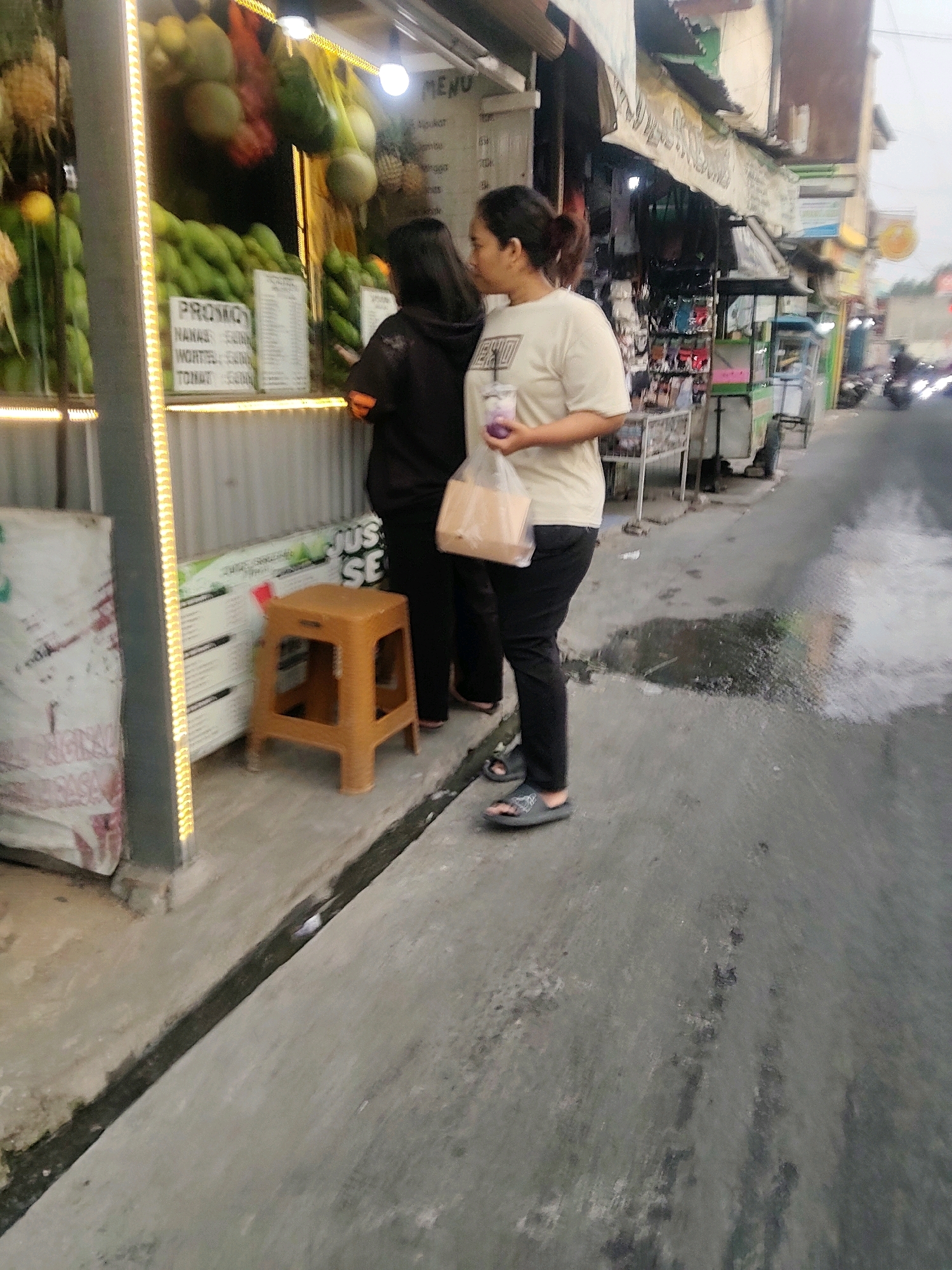 In a street-level view of a marketplace in Mekarmukti, Indonesia, two women are standing by a fruit stall. The woman on the left, wearing a dark hooded jacket and black pants, stands with her back to the viewer, facing the stall. She appears to be interacting with a menu or display. The woman on the right, wearing a light-colored t-shirt and black pants, is facing left, holding a plastic cup with a purple beverage and a plastic bag with a light brown object inside. She is wearing dark sandals. In front of them is a small, orange plastic stool. The fruit stall is brightly lit with yellow LED lights and displays a variety of fruits, including pineapples, avocados, and other green produce. Signs with text are visible on the stall. The ground is asphalt with a drainage ditch running along the edge. In the background, there are other market stalls and the blurry motion of passing vehicles, suggesting a busy urban environment.