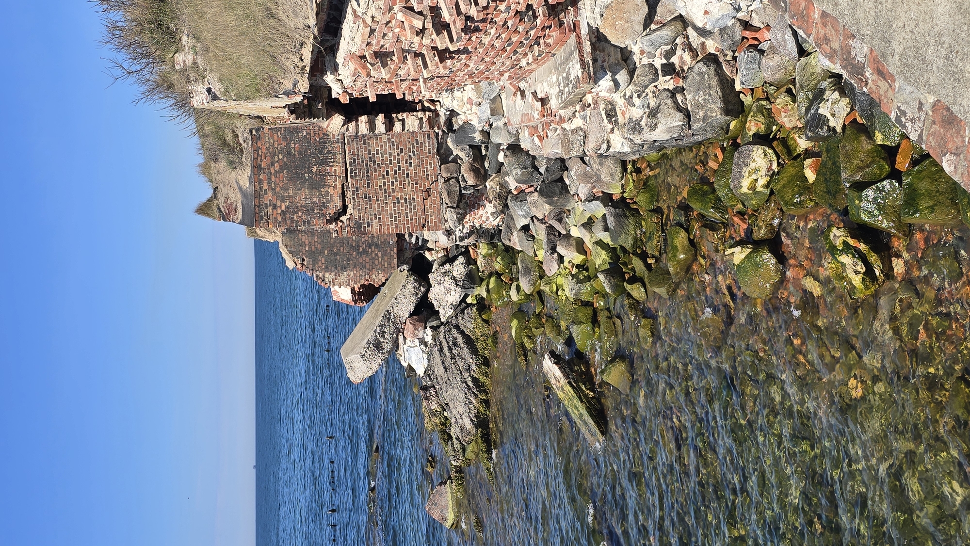 The image shows the ruins of a brick and stone structure on a coastline in Baltiysk, Russia. The structure is crumbling, with bricks and stones scattered along the shoreline. Water, appearing moss-covered, laps against the debris. The sea extends into the distance under a clear, blue sky. Vegetation is visible on the upper bank above the ruins.