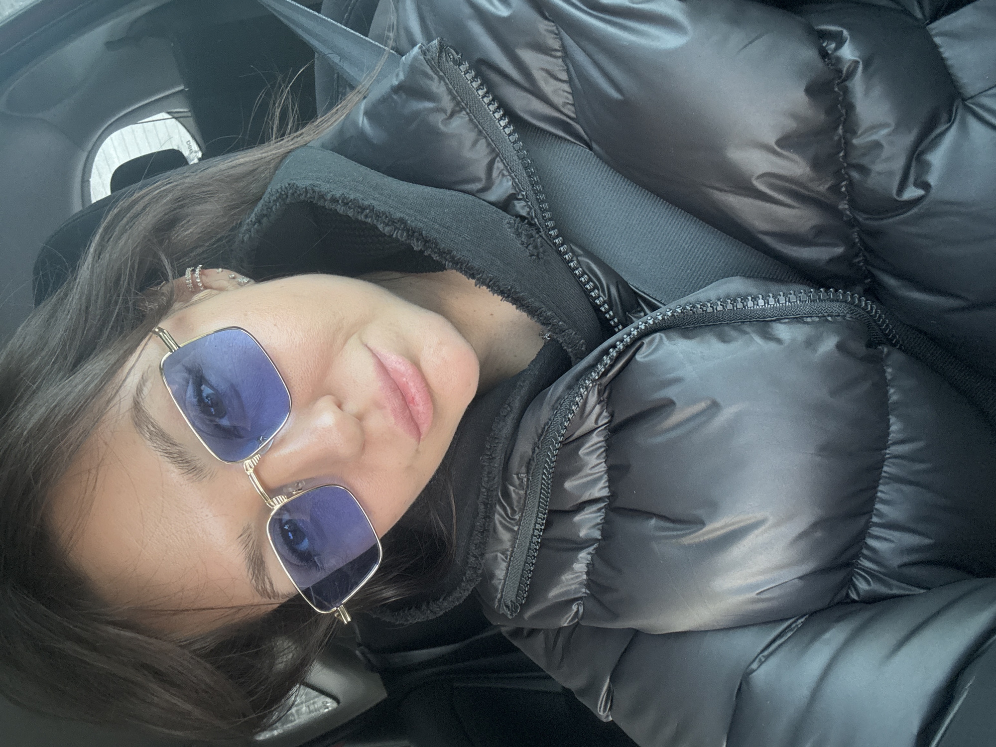 A close-up, self-portrait style photograph shows a female individual seated within the dark interior of a vehicle. The exact location in Krasnoyarsk, Russia, cannot be determined from the visual evidence. The subject, an adult with dark, straight hair, looks directly towards the camera. She wears rectangular sunglasses with gold-toned frames and blue-tinted lenses. Her lips are slightly parted. On her visible right ear, multiple earrings are present, including two stacked, paved cuff earrings on the upper helix and several smaller stud-like earrings along the lobe. She is dressed in a black, shiny puffer jacket, zipped up, over a black hooded garment featuring a visible textured or sherpa-lined collar. A black seatbelt is diagonally secured across her chest and right shoulder. A dark headrest is visible behind her right shoulder, and a blurred, light-toned background, possibly an exterior view or frosted window, is seen through a rear or side window of the vehicle.