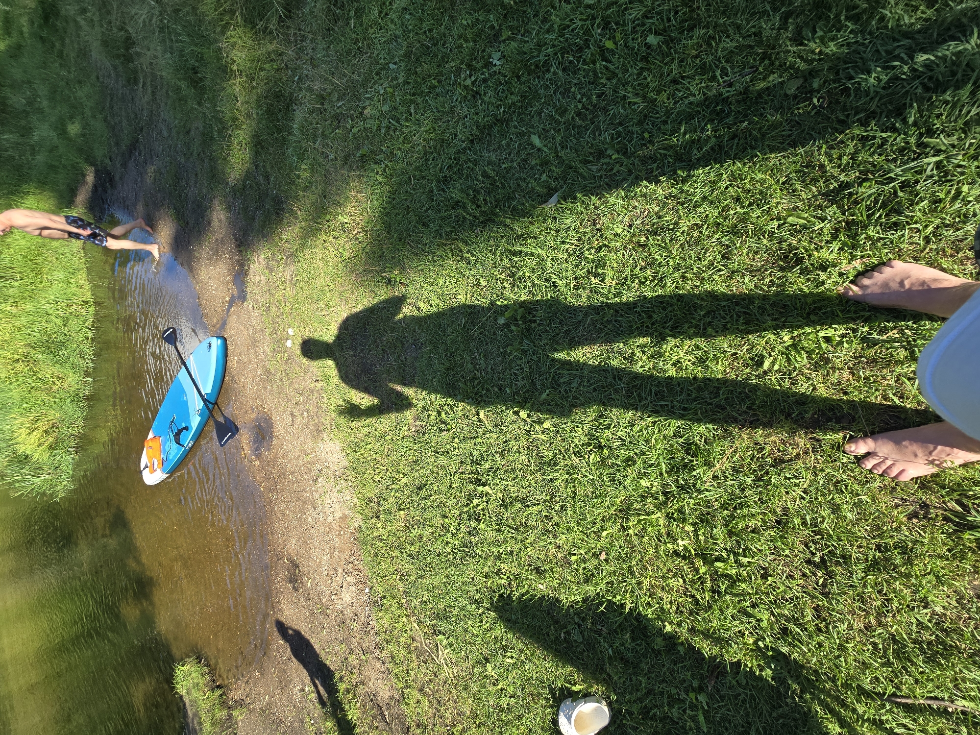 The image depicts a natural setting near a body of water. In the foreground, a person's bare feet are visible on a grassy area. A long shadow of this person extends towards the water. To the left, a white object, possibly a shoe or cup, is on the grass. Further into the scene, a blue paddleboard with an orange life vest and a paddle is floating in the shallow, clear water near a sandy bank. A second person, shirtless and wearing dark shorts, is standing near the edge of the water, with one foot in the water, and is looking down. Tall green grass lines the bank of the body of water, which appears to be a river or lake. The overall lighting suggests it is a sunny day, casting distinct shadows. The location is identified as Voinskaya Chast' Mchs, Russia.
