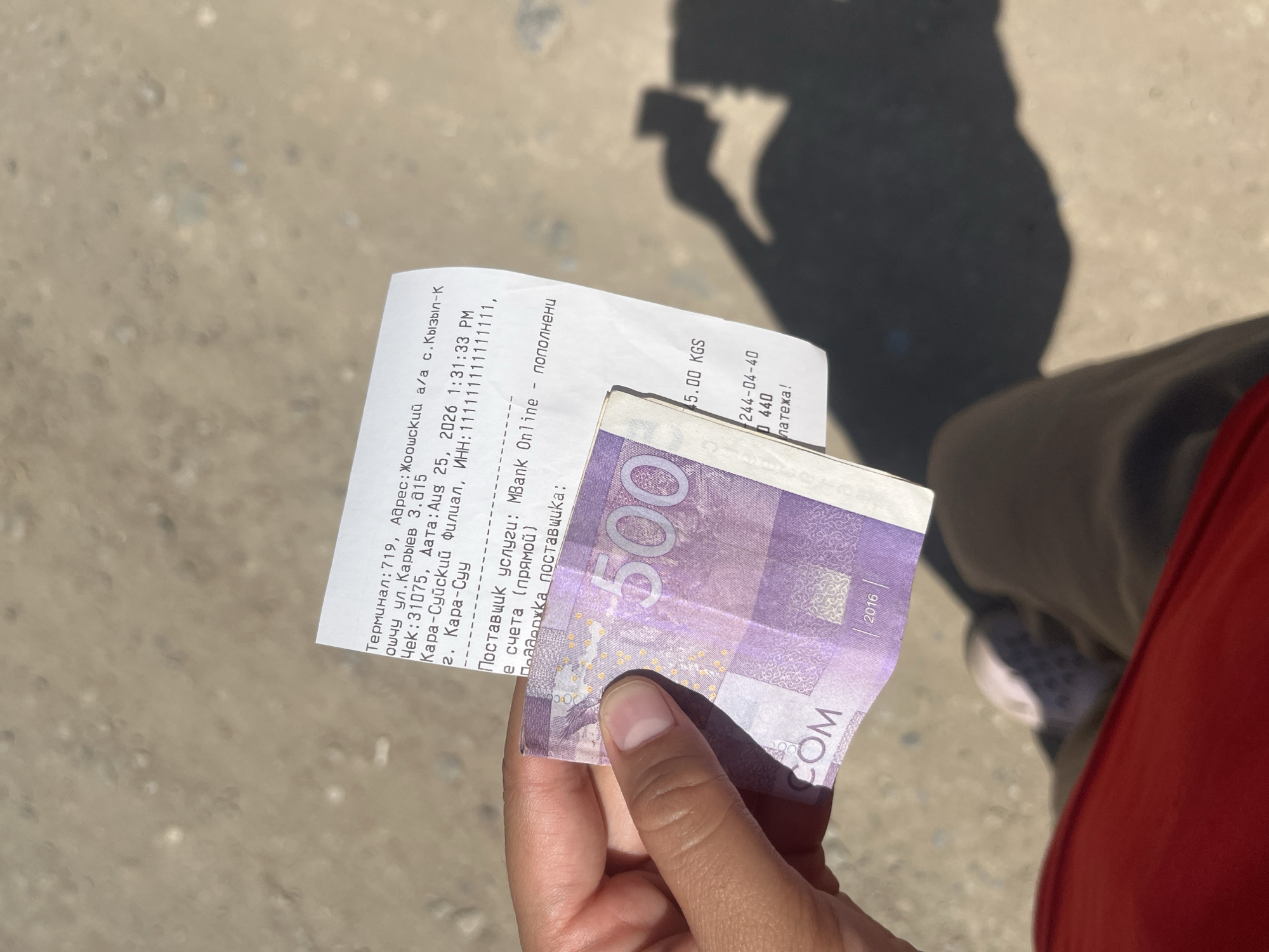 A receipt and a 500 KGS banknote are held in a person's left hand. The receipt is partially obscured by the banknote. The text on the receipt indicates it is for a terminal, address, date and time, and a payment service. The date on the receipt is August 25, 2026. The banknote is purple and features the number 500 prominently. The background appears to be an outdoor, paved surface with shadows cast from an unseen object. The setting is likely a city given the information on the receipt.