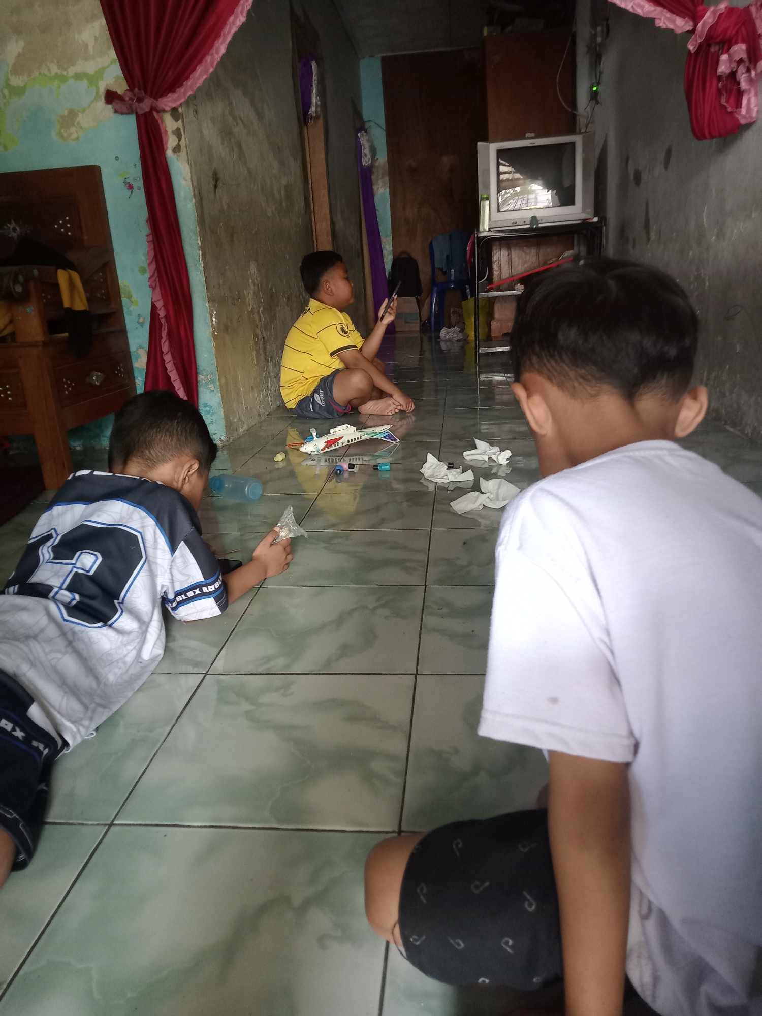 The image captures an indoor scene in what appears to be a home in Bungur, Indonesia. The floor is tiled with a green and white pattern. Three young boys are present. One is lying on his stomach, wearing a white and blue Roblox-themed shirt with the number 2 on the back, and is holding a small plastic wrapper in his hand. Another boy is sitting cross-legged, wearing a yellow striped shirt and shorts, holding a phone. The third boy is facing away from the camera, wearing a white T-shirt and black shorts. Scattered on the floor around the boys are toys, including what appears to be a toy helicopter, a plastic bottle, and crumpled pieces of tissue. In the background is a wall with peeling paint, adorned with red and pink curtains. A television set is placed on a stand.
