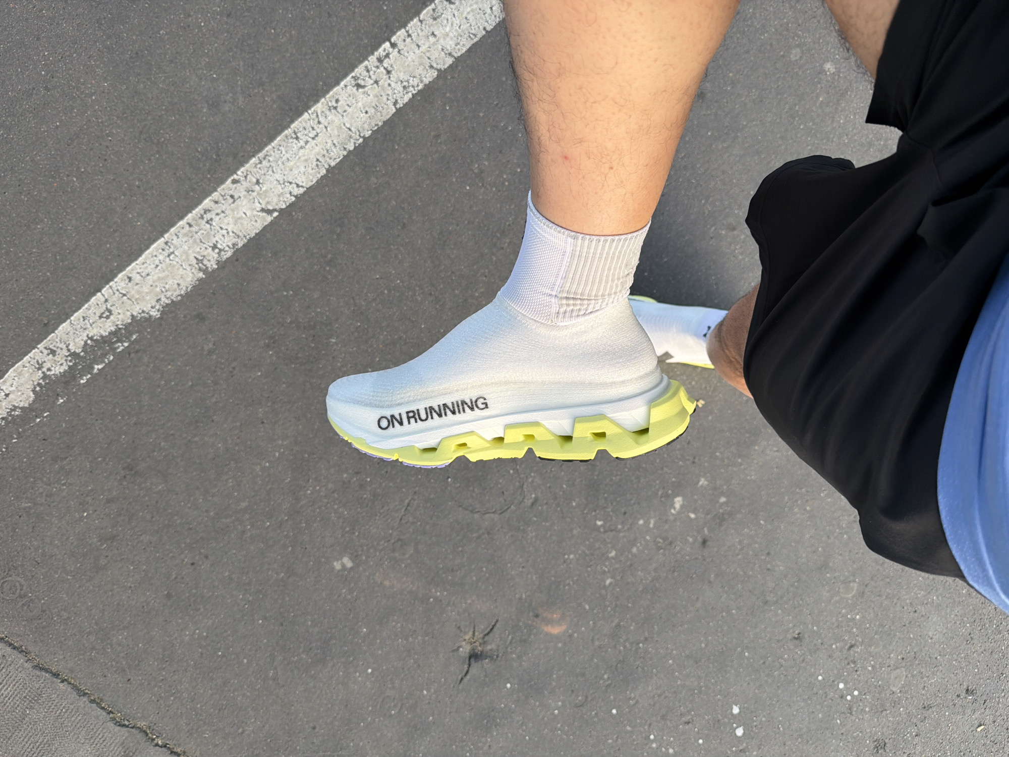 The image shows a person standing on asphalt, with a partial view of a white “On Running” brand shoe with a yellow sole, a white sock, a bare leg, and black shorts. A white painted line is visible in the upper left corner of the frame. There's also a spider on the ground near the shoe. The location appears to be an outdoor paved area, potentially a parking lot or a street in an urban environment. There is no evidence to indicate that the location is Paris, France.