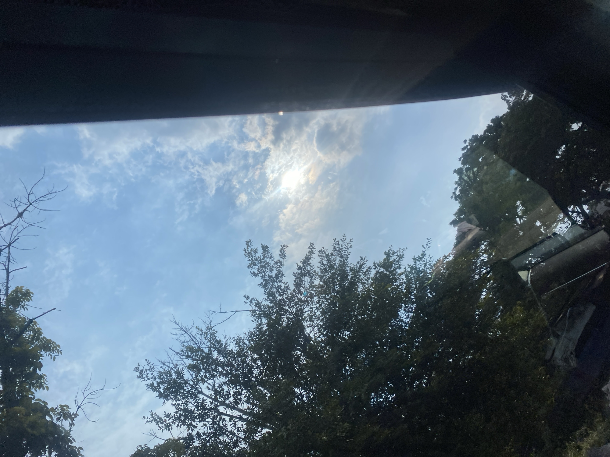 The image shows a view looking upwards from inside a vehicle, with a large portion of the frame dominated by a bright blue sky filled with scattered white clouds. Sunlight pierces through the clouds in the upper right quadrant, creating a lens flare effect. To the left, the dark green foliage of trees frames the sky, with some bare branches extending into the blue. A dark, solid object, likely part of the vehicle's interior, occupies the right side of the image, obscuring a portion of the view. The reflections on the vehicle's windows show more trees and sky. The image appears to be taken during the daytime in an outdoor environment, possibly with a focus on the natural elements framed by the vehicle. No specific landmarks or signs of the city of Gambut, Indonesia, are discernible.