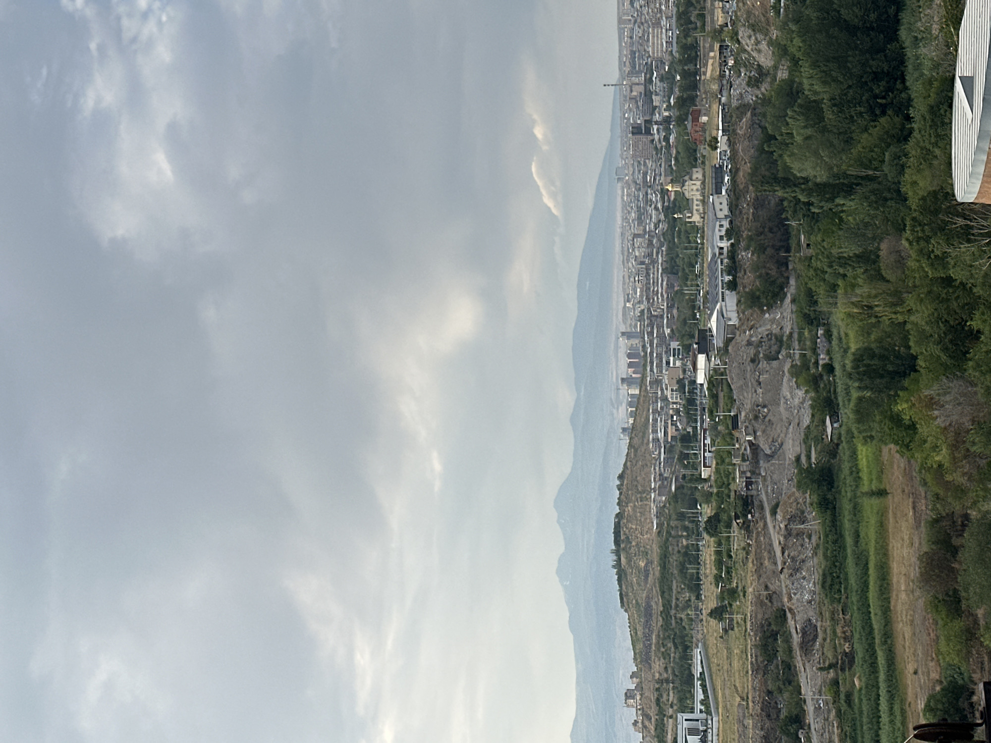 This is an aerial, eye-level view of the city of Yerevan, Armenia, looking towards the mountains under a cloudy sky. In the foreground, there is a terraced hillside with sparse vegetation, rubble, and some small structures. Further back, the city spreads out with numerous buildings, a prominent church with a golden dome, and industrial structures. In the distance, a range of mountains forms the horizon, appearing hazy and light blue. The overall impression is of a sprawling urban landscape set against a natural backdrop.