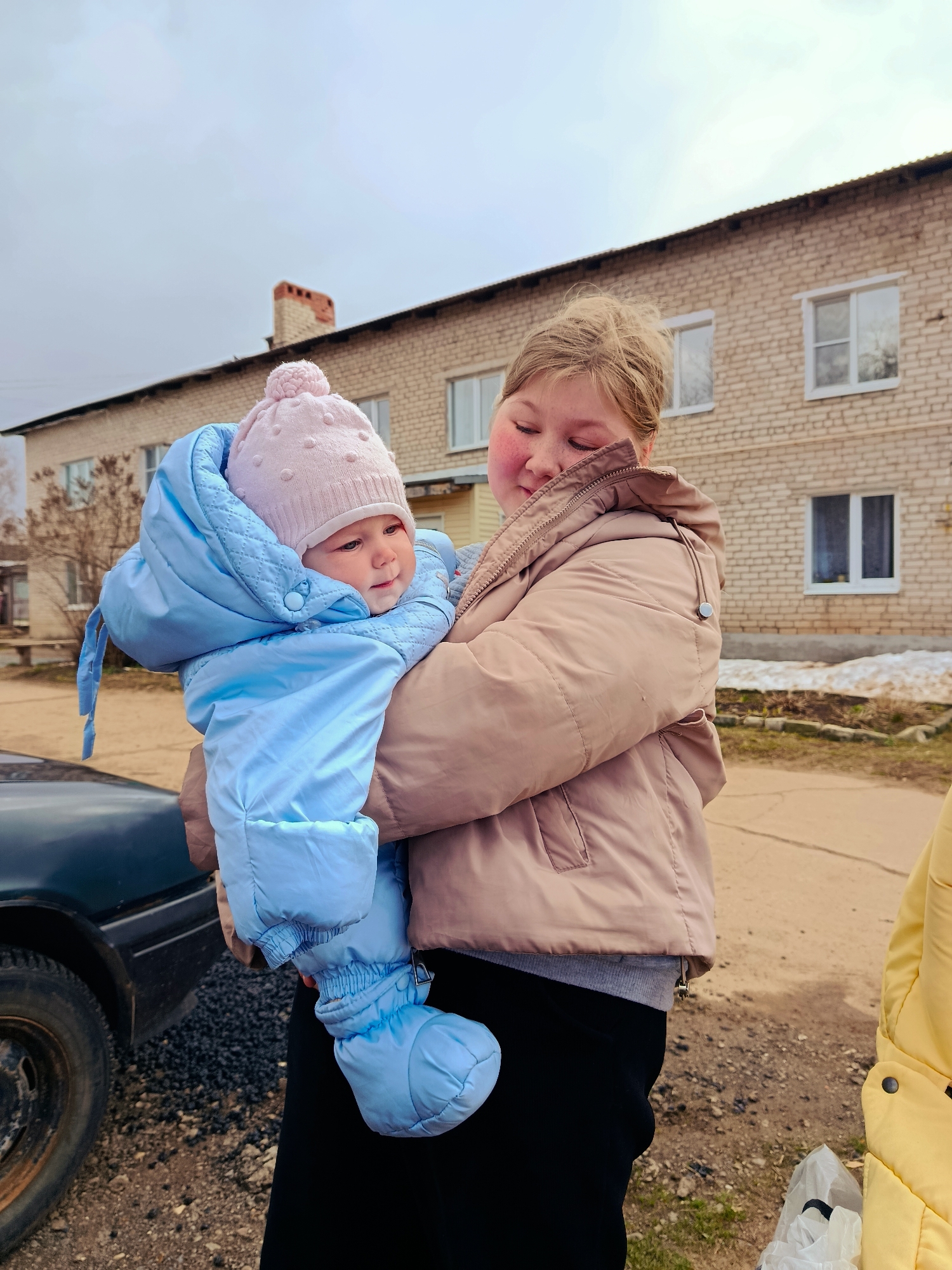 An outdoor image from Fokino, Russia, captures a tender moment between a young woman and a baby on an overcast day. The young woman, with light hair tied up and flushed cheeks, wears a light brown puffer jacket, a grey top, and black pants. She holds the baby close to her, with her eyes softly closed or looking down, conveying a sense of affection or gentle comfort.
The baby is dressed warmly in a light blue quilted snowsuit or bunting bag, complete with matching booties and a light pink knitted hat featuring a pom-pom. The baby's face is visible, looking towards the right with a neutral or slightly curious expression.
In the background, a two-story brick building with light-colored bricks and white-framed windows is visible, suggesting a residential or apartment setting. A red brick chimney is prominent on the roof. The ground is a mix of dirt and gravel, with patches of melting snow indicating cold weather, likely late winter or early spring. A dark-colored car is partially visible on the far left, and a white plastic bag lies on the ground to the far right. The overall scene suggests a casual moment outdoors in a somewhat utilitarian or residential environment. No visible text is present in the image.