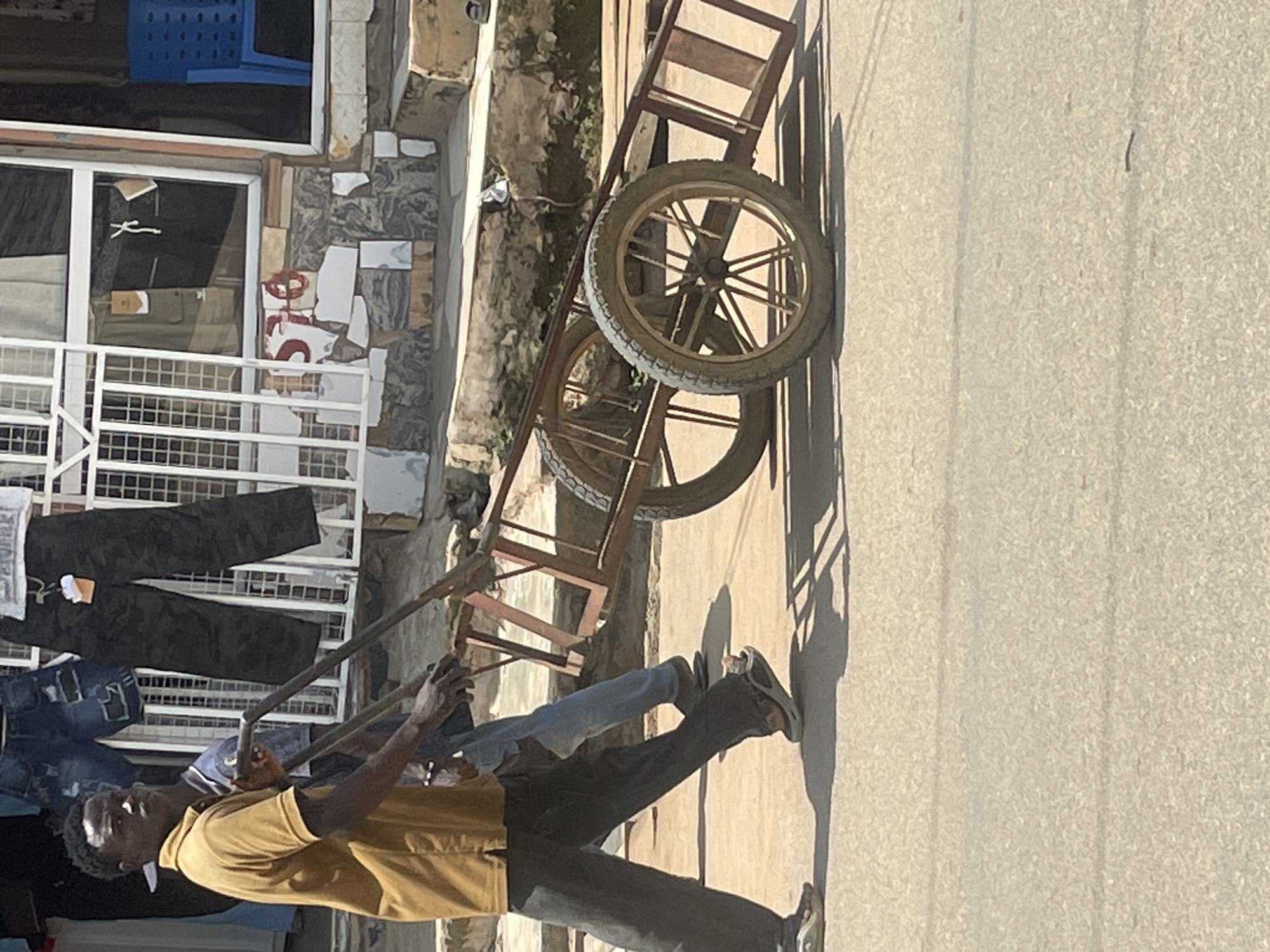 A young man in a tan polo shirt and dark pants is pushing a two-wheeled cart with a metal frame and wooden slats in front of a shop. The cart has two large spoked wheels, one of which is clearly visible. Behind the man, another person in a blue shirt and jeans is walking alongside the cart, appearing to assist. The shop behind them displays clothing, including jeans and dark camouflage pants, hanging on a rack. A blue plastic chair is visible through a window. The ground is paved, and the scene appears to be outdoors on a street in Abuja, Nigeria.