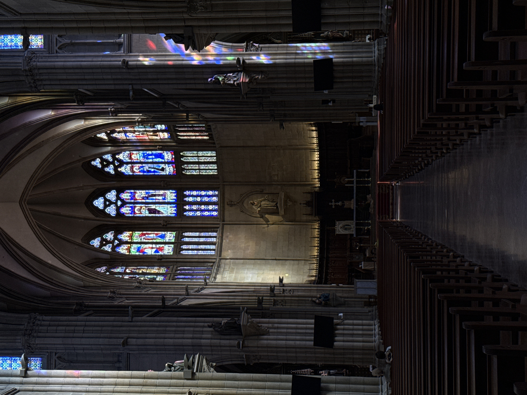 The image depicts the interior of a grand Gothic cathedral in San Sebastián, Spain. The scene is dominated by rows of dark wooden pews, stretching into the distance, with a polished floor reflecting the ambient light. Ornate stained-glass windows, featuring vibrant blue and red hues, illuminate the interior with colorful light, casting intricate patterns on the stone columns and walls. Statues of religious figures are visible on pedestals along the columns. In the foreground, two individuals are seated on the pews, facing the altar area, which is partially obscured by the angle of the shot. The overall atmosphere is solemn and reverent, with the architecture of the cathedral creating a sense of awe and history.