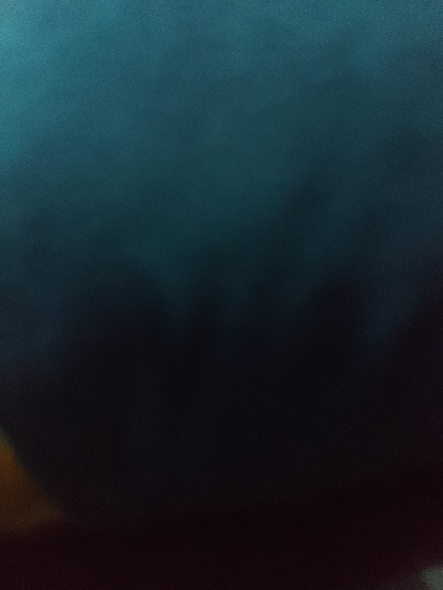 The image is a dark, abstract composition dominated by shades of deep blue and black. A subtle gradient transitions from a lighter teal or turquoise at the top to a near-black at the bottom. There is a grainy texture throughout the image, suggesting low light conditions or a deliberate stylistic choice. In the bottom left corner, a faint, diffused area of orange or yellow light is visible, contrasting with the predominantly cool tones. No discernible people, subjects, objects, or actions can be identified due to the abstract and blurred nature of the image. The scene appears to be devoid of any specific environmental markers that would place it within the city of Jauro Yinu, Nigeria, or any other recognizable location.
