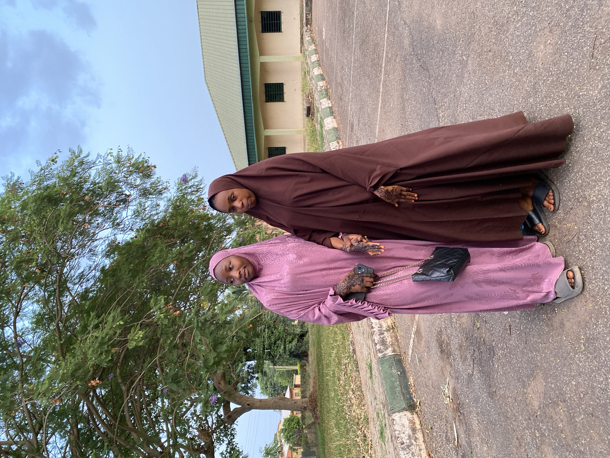 Two women wearing hijabs stand side-by-side on a paved surface, likely a street or walkway in Vom, Nigeria. One woman is wearing a brown hijab, and the other is wearing a purple hijab and is holding a black quilted handbag on a gold chain. Both have henna tattoos on their hands. The area appears to be a university campus with a building and a tree in the background.