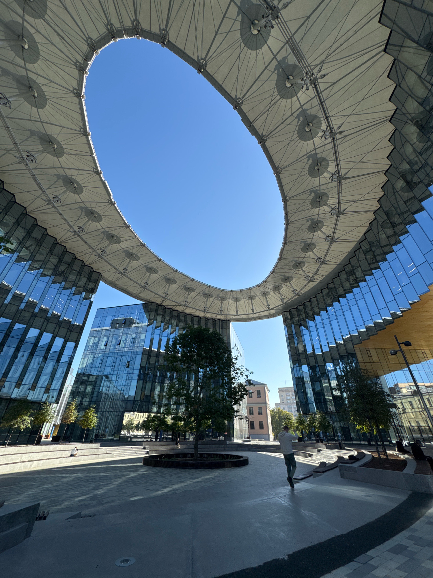 The image captures a low-angle view of the modern architectural design of the Central Children's Store on Lubyanka Square in Moscow, Russia. The iconic oval-shaped roof structure, composed of a translucent white membrane supported by a complex metal framework, dominates the upper portion of the frame, revealing a clear blue sky through its opening. Beneath this canopy, sleek, glass-fronted buildings with reflective facades rise up, their surfaces mirroring the sky and surrounding structures.
In the foreground, a paved public space features tiered seating areas and planters with young trees. A lone individual, wearing a light-colored hoodie and dark pants, is seen walking across the open area, their back to the camera. Other individuals are seated on benches in the background, engaged in passive activities. The scene is bathed in bright sunlight, casting distinct shadows on the ground, indicating a clear day. The overall impression is one of contemporary urban design and public space utilization.