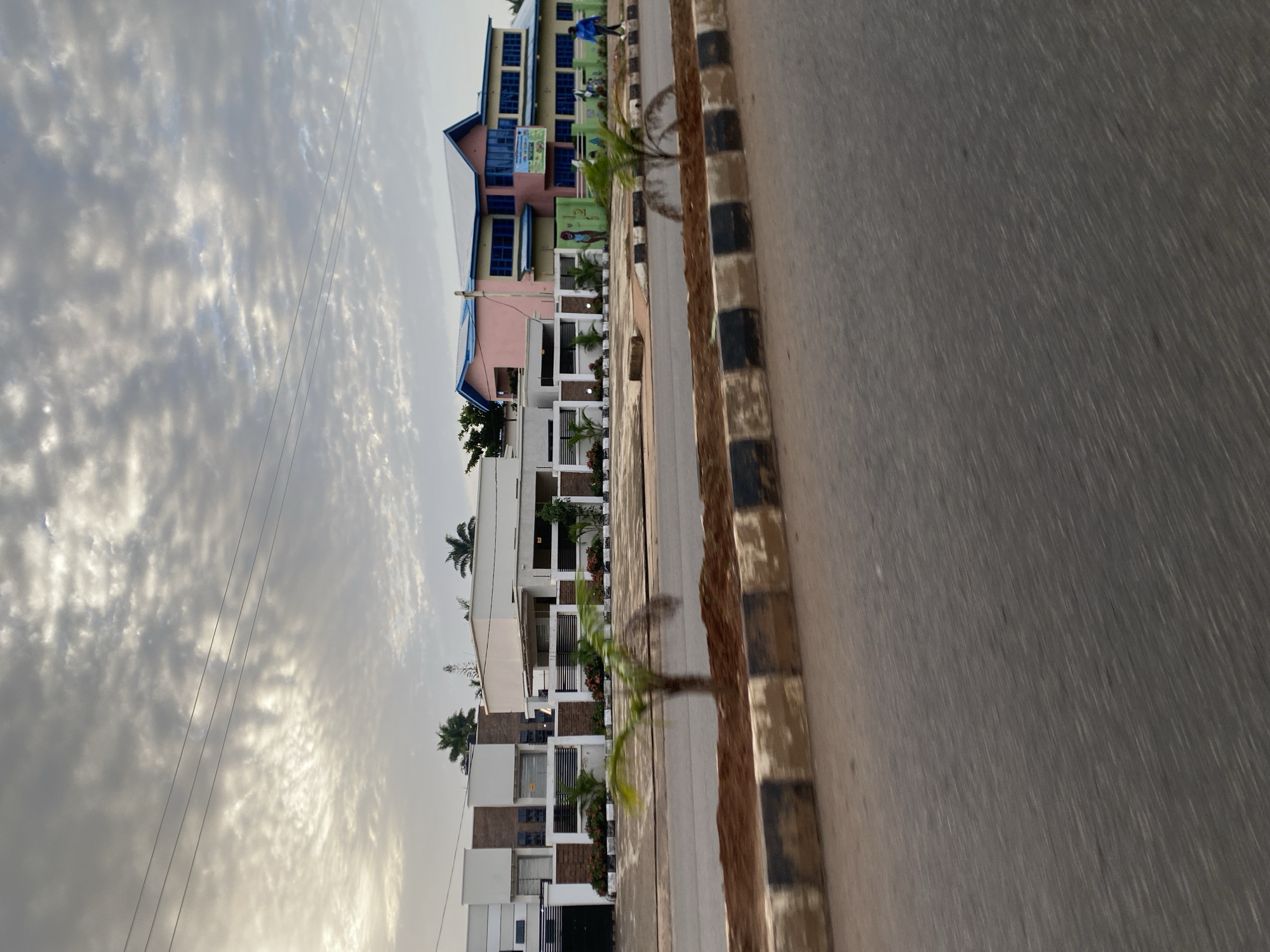 This is a photograph of a street scene in the city of Bar Jirgi Summa, Nigeria. In the background, there are several buildings. The building on the right is a multi-story structure with a pink and yellow facade and blue window frames. It appears to be a school or educational institution, as indicated by a sign on its side with colorful graphics. To the left of this building is a modern, single-story white building with a fence. Further left, another section of a white building is visible.
The foreground features a street with a paved surface, appearing slightly blurred due to motion. A median strip runs along the street, bordered by concrete structures with black and white stripes. This median is covered in dark soil and contains several small palm trees. Overhead, power lines stretch across the frame against a cloudy sky. A single person in a blue jacket is walking on the sidewalk in front of the school building. The overall impression is that of a well-maintained street and buildings on a somewhat overcast day.