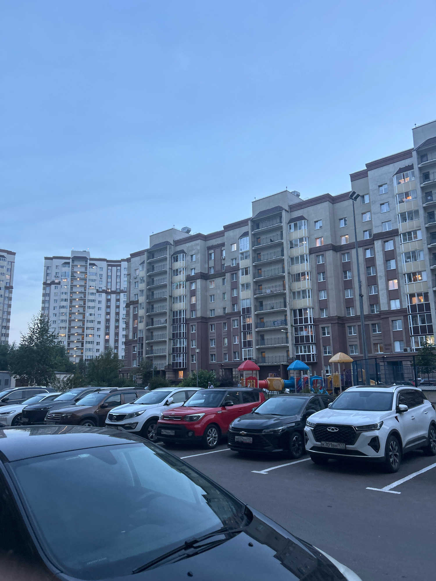 An outdoor, eye-level, wide shot captures a parking lot in front of residential buildings in Domodedovo, Russia. The sky is overcast, and the lighting suggests dusk. Several cars are parked in a designated lot. The buildings, tall and modern, feature balconies and numerous windows, some of which are illuminated. A playground with colorful structures is visible in front of the buildings. The scene is orderly and depicts a typical urban residential environment.