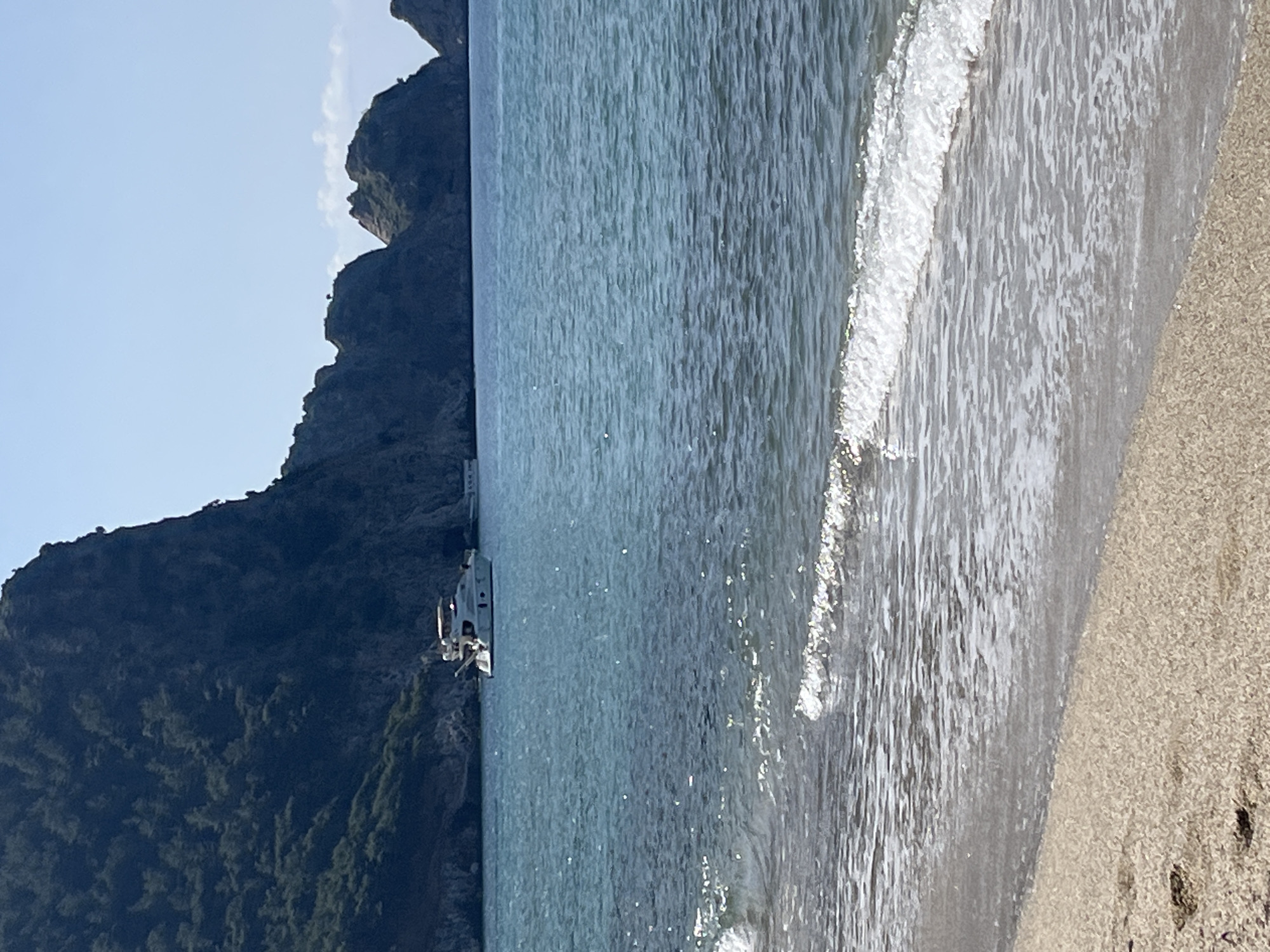 The image depicts a coastal scene in Alanya, Turkey. A sandy beach occupies the foreground with visible footprints. Waves are gently lapping onto the shore. In the midground, the sea extends towards a mountainous coastline. A yacht is anchored near the coast. The sky is clear with a blue hue. The mountain slopes are covered in green vegetation.