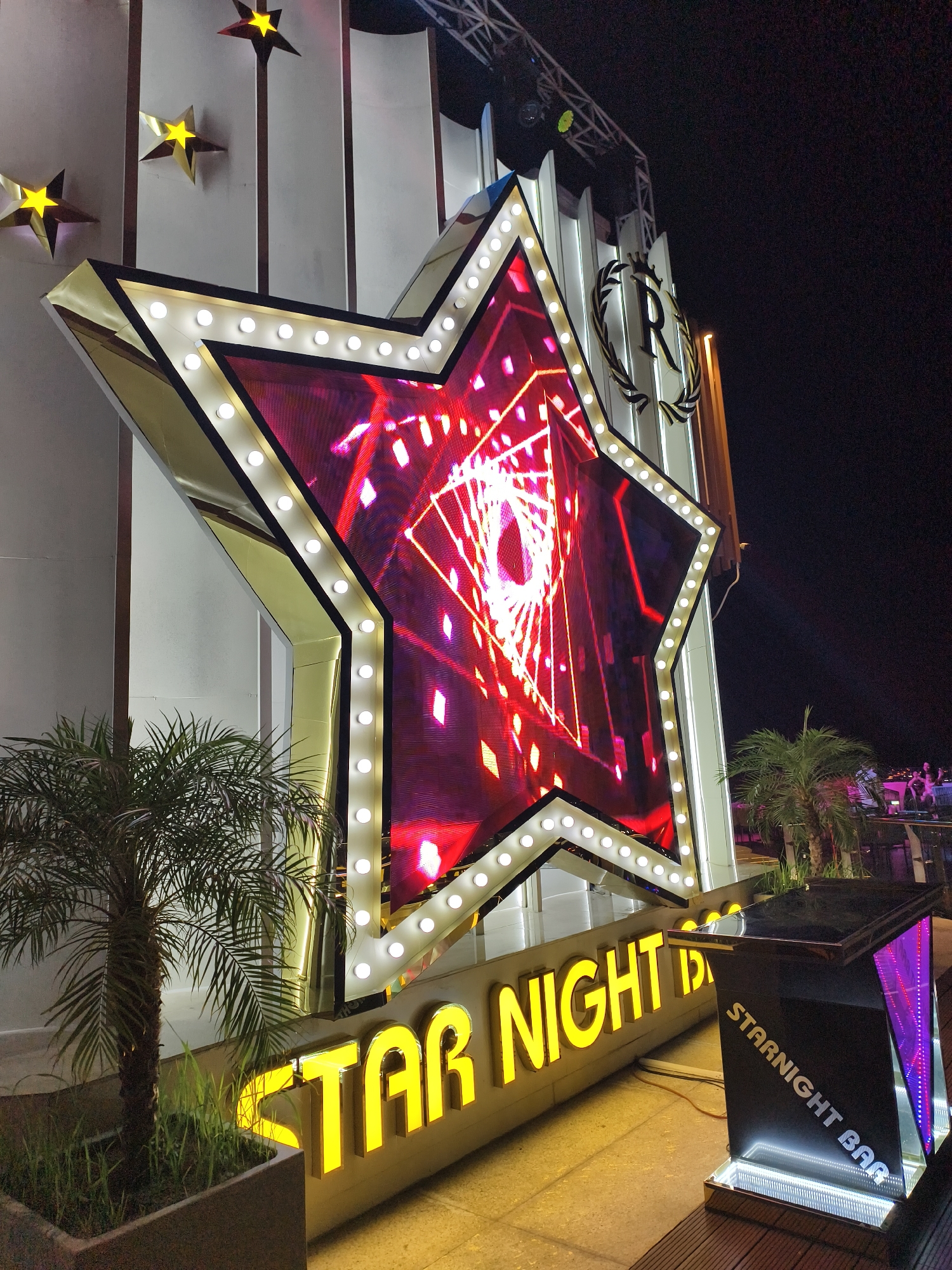 This nighttime photograph captures a vibrant outdoor entertainment venue, likely a bar or club, in Nha Trang Ward, Vietnam. The central focus is a large, illuminated five-pointed star structure featuring an LED screen that displays dynamic, abstract patterns in red and white. The perimeter of this star is adorned with numerous bright white circular lights, evoking a classic marquee style.
Below the large star, prominently displayed in bright yellow, illuminated letters, is the text "STAR NIGHT". To the right, a sleek, modern bar stand is visible, featuring reflective black surfaces and internal purple and pink lighting. The words "STARNIGHT BAR" are written vertically in white on its side.
Small palm trees in dark planters are positioned on either side of the main star structure, adding a tropical element to the scene. In the upper left background, several smaller, three-dimensional golden stars are mounted on a white, vertically ribbed facade. Higher up on this facade, a distinct logo appears, consisting of a stylized 'R' topped with a crown and encircled by a wreath. Metal truss structures, likely for stage lighting or sound equipment, are also visible against the dark night sky. In the distant background, indistinct figures of people can be seen on what appears to be an elevated platform, suggesting a lively social gathering. The abundance of bright, artificial lights creates a festive and energetic atmosphere typical of an evening entertainment spot.