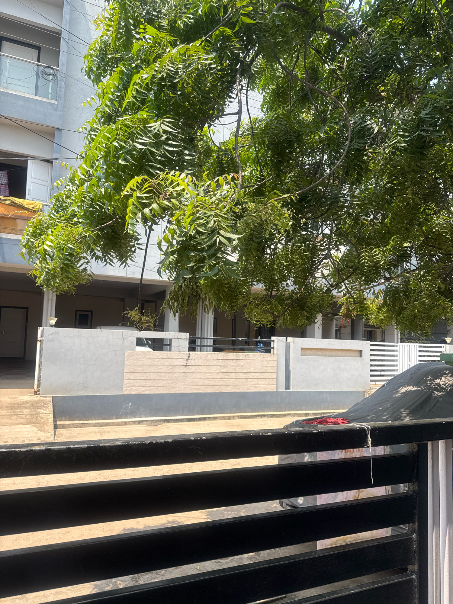 The image shows a view of a residential building and its surroundings in Nagamalli Thota, India. A large tree with dense green foliage dominates the upper portion of the frame, casting shadows on the scene below. In the background, a multi-story building with a light gray facade is visible. The building has several windows and balconies, some with glass railings. Below the building, there is a covered parking area or ground floor with pillars and doors. A concrete wall with horizontal slats separates the building area from the foreground. In the immediate foreground, a black metal gate with horizontal bars is prominent. A vehicle covered with a dark tarp is parked behind the gate. A small red cloth item is tied to the top bar of the gate with a white string. The ground appears to be paved or concrete. The scene suggests a bright, sunny day.