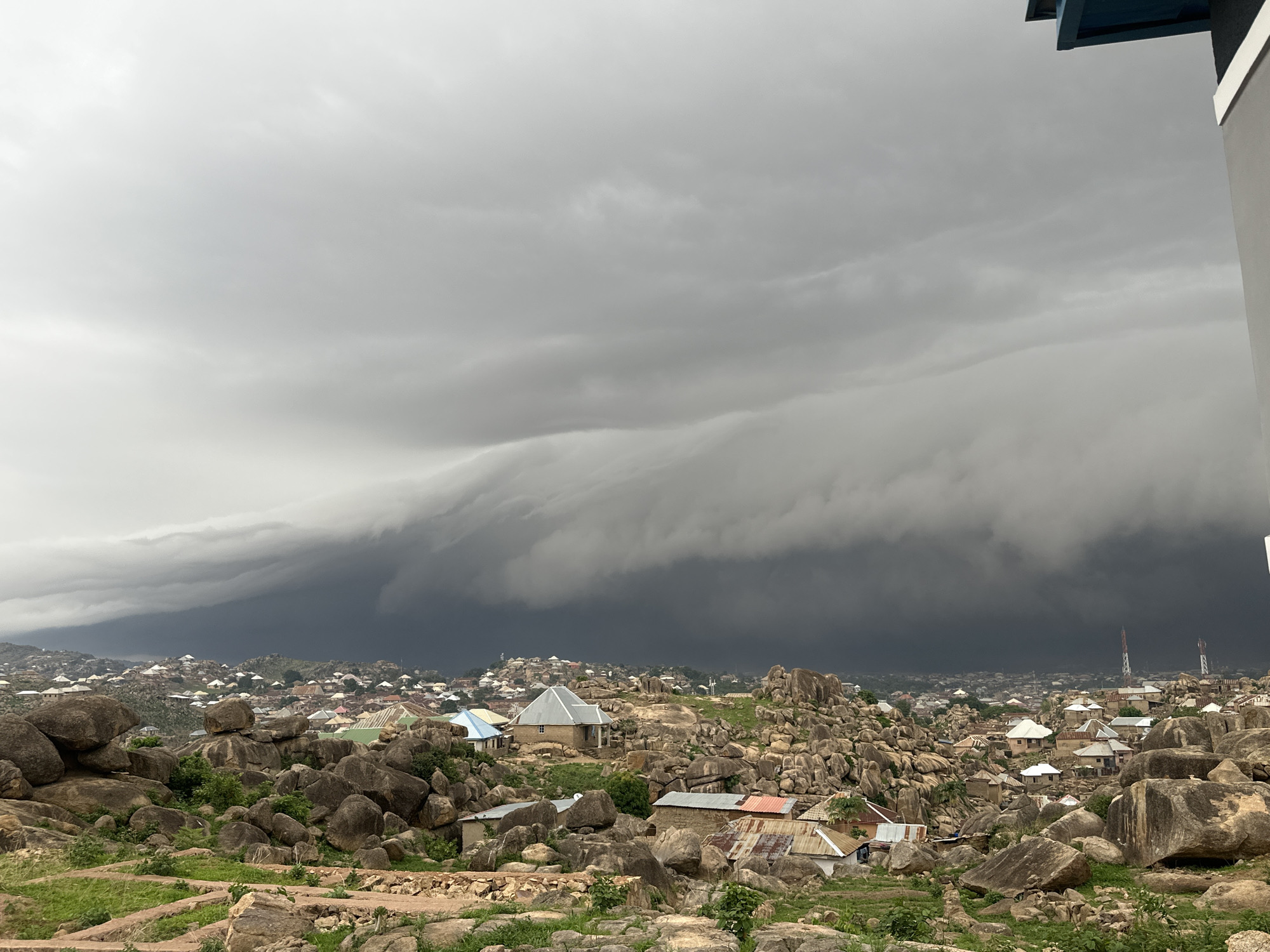 The image captures a striking storm front rolling in over a section of Jos, Nigeria. The foreground features rocky terrain with scattered vegetation, and small, simple houses are nestled among the rocks. The town extends into the distance. Dominating the sky is a thick, ominous band of dark storm clouds, indicating a severe weather event approaching.