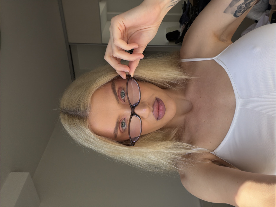 This image captures a frontal, head-and-upper-torso view of a single adult female subject indoors in Budva, Montenegro. The subject has fair skin and long, straight blonde hair parted centrally. She wears a white spaghetti-strap camisole with a ribbed texture on the chest. Her eyes, appearing blue-green, are directed towards the camera. Her face features pink eyeshadow and matte mauve lipstick. Her right hand is holding a pair of dark-framed eyeglasses by the temple, positioned just below her eye line. A tattoo, depicting a stylized figure or face, is visible on her left upper arm. The background on the left consists of a plain, light-colored wall, while the right side reveals a partially open mirrored wardrobe containing folded clothing. Natural light appears to be entering from the left side of the frame.