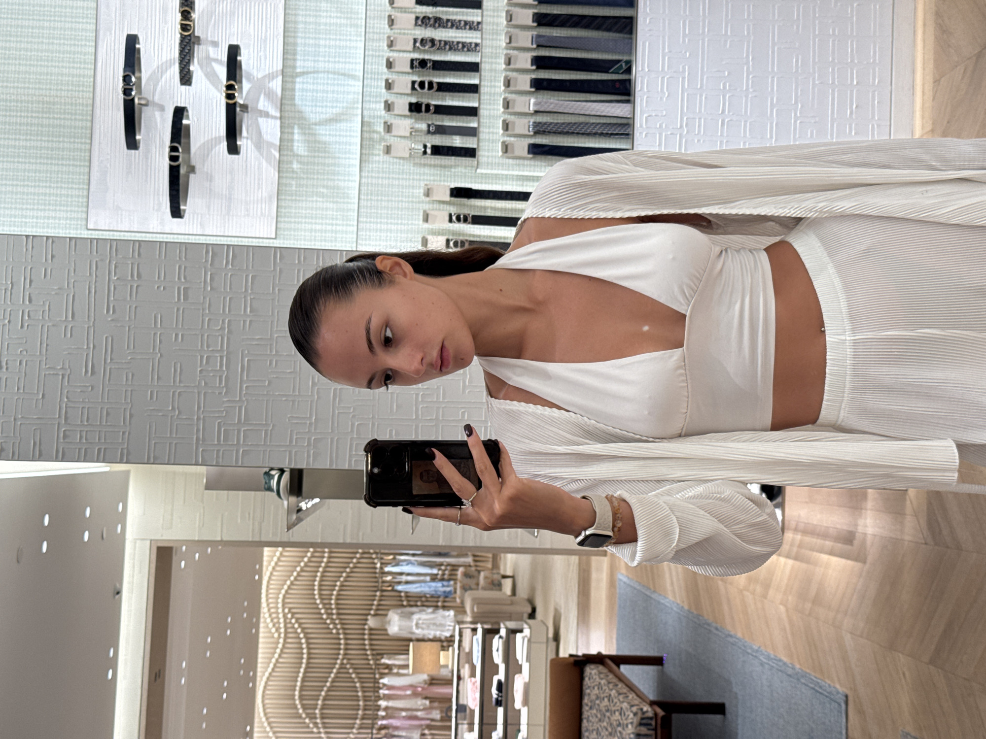 A woman is taking a selfie in what appears to be a high-end retail store. The store features displays of belts and bracelets. The woman is wearing a white outfit. The location is possibly in the city of Wichit, Thailand.