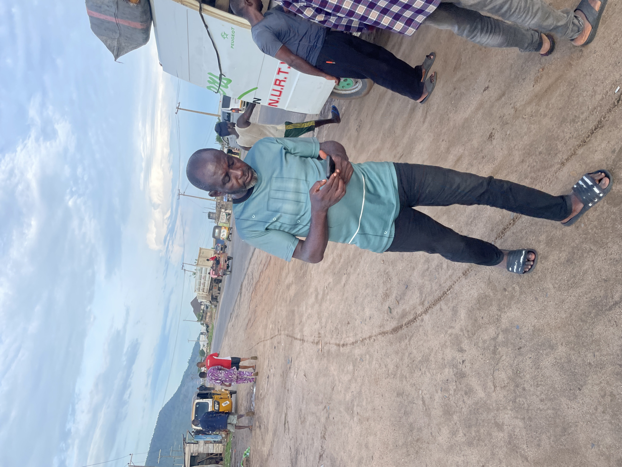 An outdoor, medium shot shows a man looking down at his phone while standing in a dirt lot in Lokoja, Nigeria. The man, wearing a teal short-sleeved shirt and black pants, has his thumbs on his phone screen. He stands in the foreground, facing right. To the right and behind him, two other men stand near a white truck with the words "N.U.R.T." and "PEUGEOT" written on its side. The truck is loaded with large, dark bags on its roof. The truck is parked on the side of a paved road that stretches into the distance. Several other vehicles, including a yellow tricycle, are visible on the road. In the background, there are buildings, power poles, and a large hill under a cloudy sky. The ground is uneven dirt with tire tracks visible.