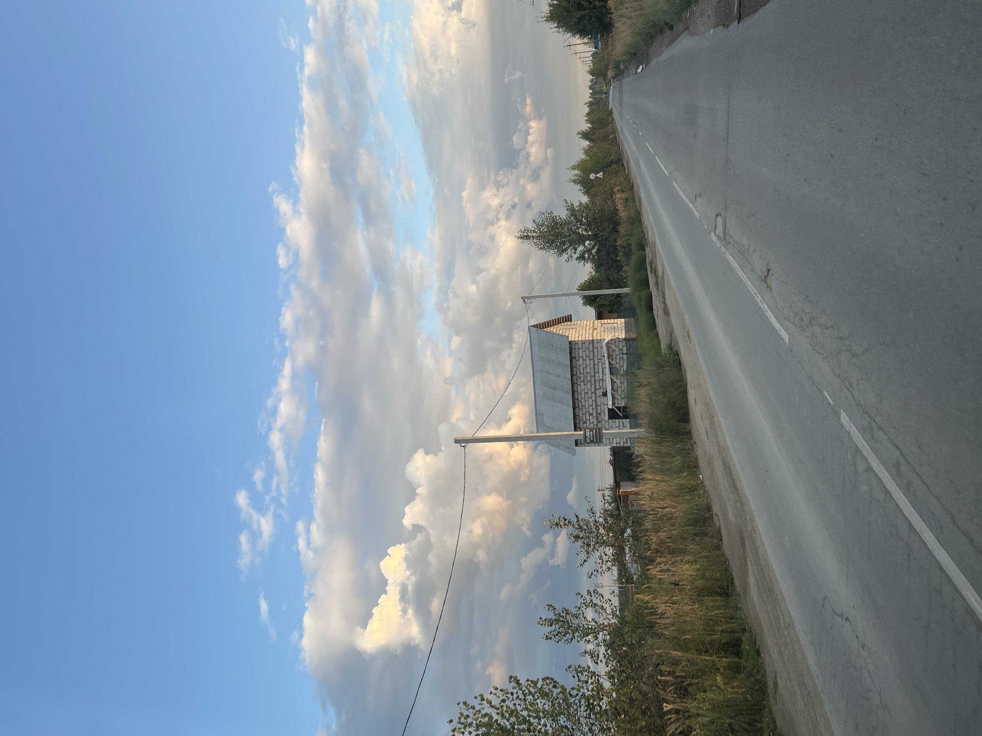 The image captures a rural landscape with a paved road stretching into the distance under a partly cloudy sky. To the left of the road, a small, unfinished white brick building with a grey metal roof is visible, situated behind a fence and surrounded by tall dry grass and scattered trees. A utility pole stands beside the building, with power lines extending across the scene. The right side of the road is bordered by a dense line of trees and vegetation. The sky features large, fluffy white clouds illuminated by sunlight, contrasting with patches of blue sky. The scene appears to be located in the village of Mochishche, Russia, given the context. There are no people or animals depicted in the image, and the overall atmosphere is tranquil and undeveloped.