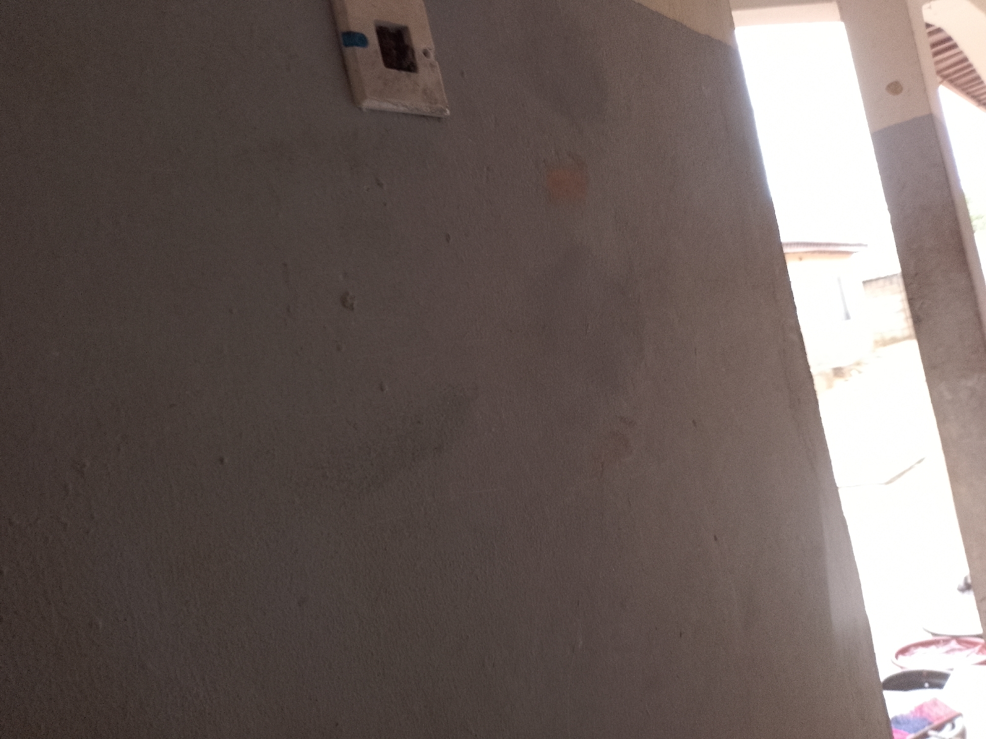 The image captures a close-up view of a textured, grey wall with a light-colored electrical outlet box mounted on its surface. The outlet box, positioned towards the top left of the frame, appears to be an older style. The wall itself exhibits some subtle imperfections, including faint water stains or discolorations, one of which is a light orangish hue.
To the right of the wall, a bright, sunlit area is visible, suggesting an outdoor or open-air setting, possibly a walkway or courtyard. Through the gap between architectural elements, hints of buildings and possibly some red-colored objects on the ground can be seen. The bright lighting suggests it is daytime, likely with clear weather. The overall impression is one of an outdoor, possibly residential or public space, with a focus on the architectural details of the wall and its immediate surroundings. No people are visible in the frame. There is no discernible text in the image.