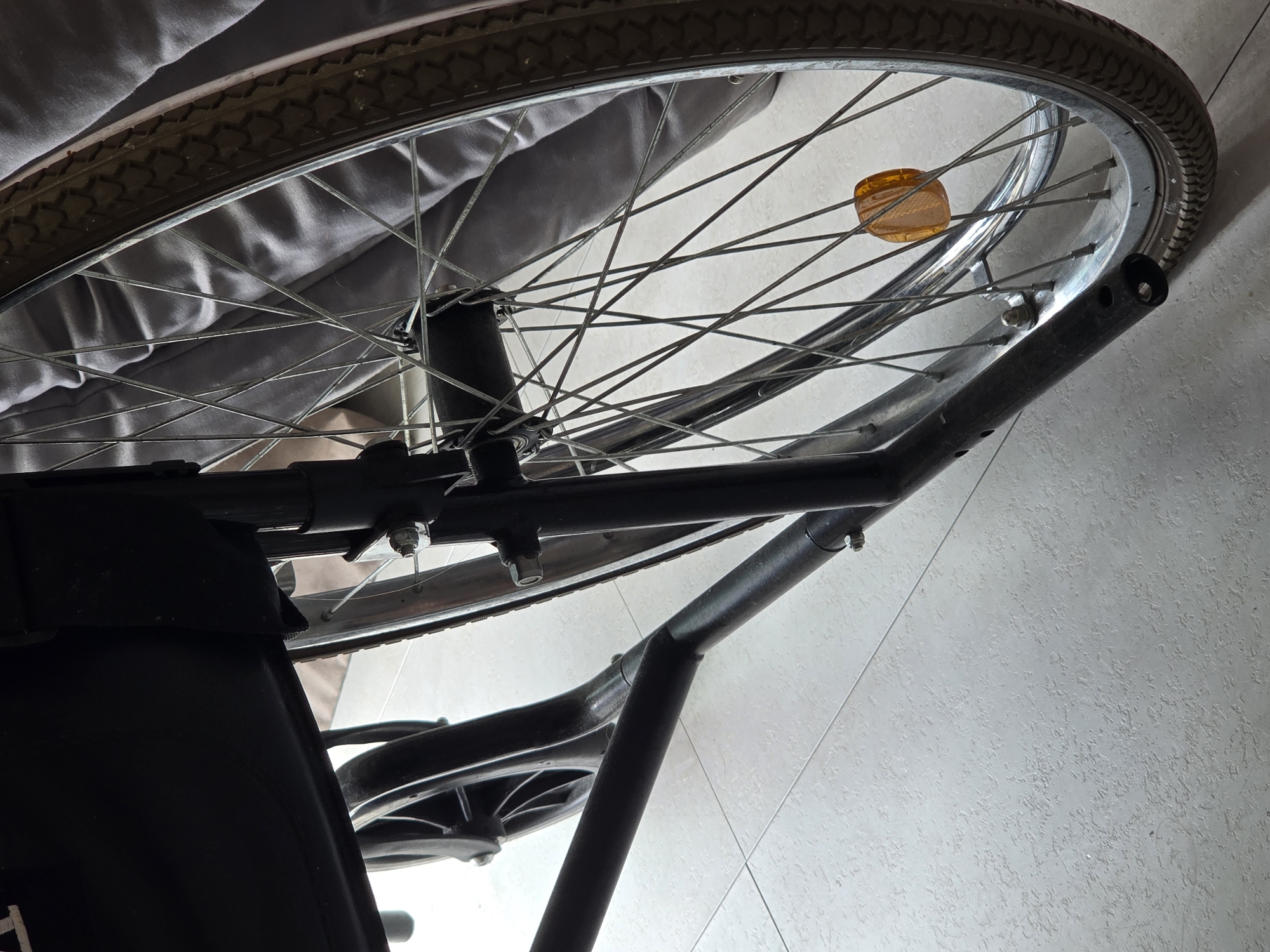 A close-up, low-angle shot focuses on the wheel and frame of a black wheelchair, resting on a light-colored tiled floor. The wheel features a silver rim and numerous thin metal spokes, with a dark-colored tire exhibiting a tread pattern. A small, orange reflector is attached to the spokes. To the left, a portion of the black fabric seat of the wheelchair is visible. The background is a soft blur of a gray, textured surface, possibly fabric or a blanket. The lighting suggests an indoor environment. The image does not provide any information about its location in Ulan-Ude, Russia, or any people or actions.