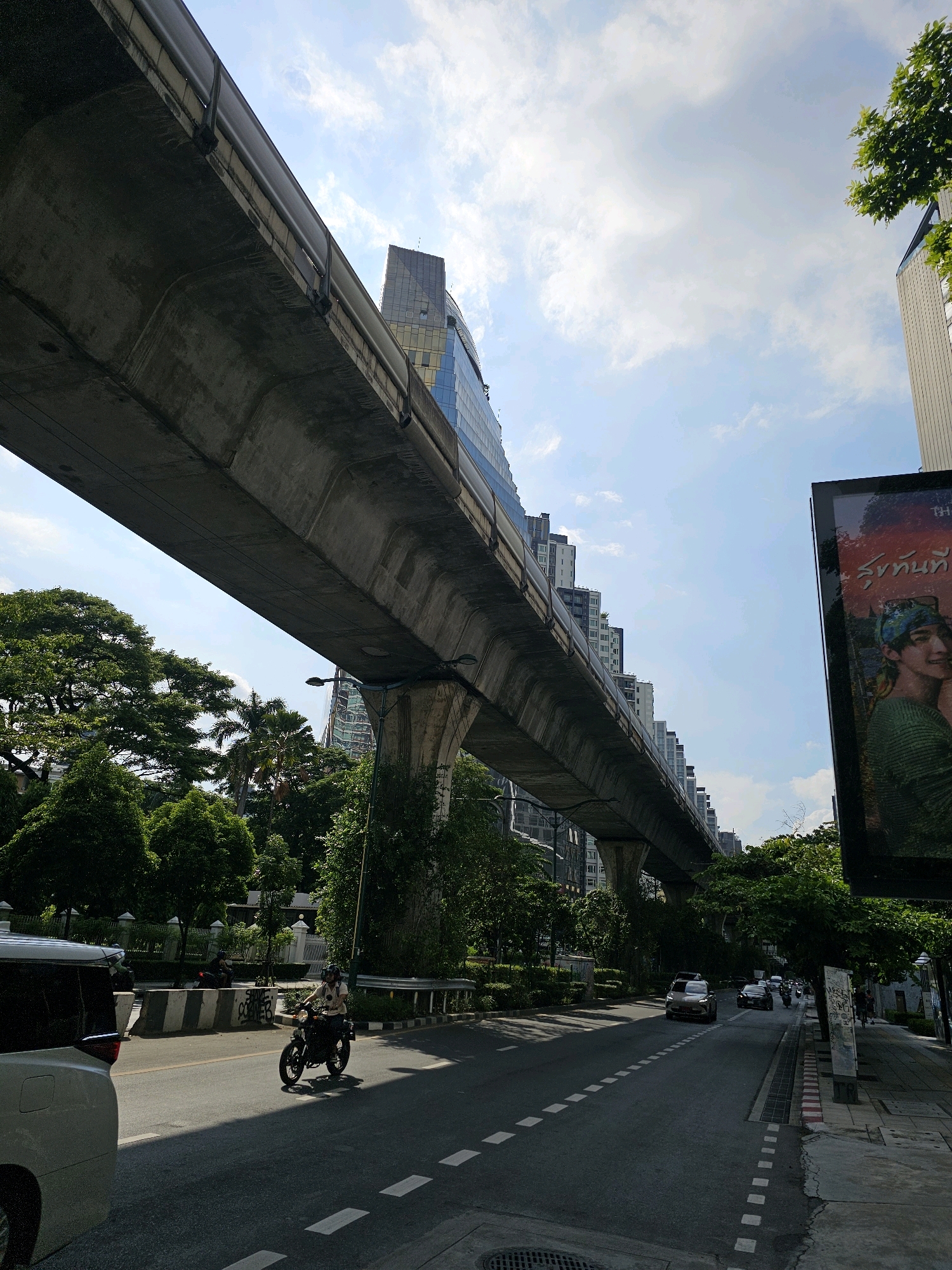 A street view in Bangkok, Thailand, features a concrete elevated train track running diagonally across the frame from left to right, casting a shadow on the road below. Underneath the track, a dense canopy of green trees lines the side of the road, with tall buildings visible in the background. A person rides a motorcycle on the asphalt road, which has white dashed lines indicating lanes. A white car is parked on the left side of the road, and several other vehicles are driving on the road further down. A large billboard with a colorful image and Thai text is positioned on the right side of the street. The sky is partly cloudy with patches of blue.