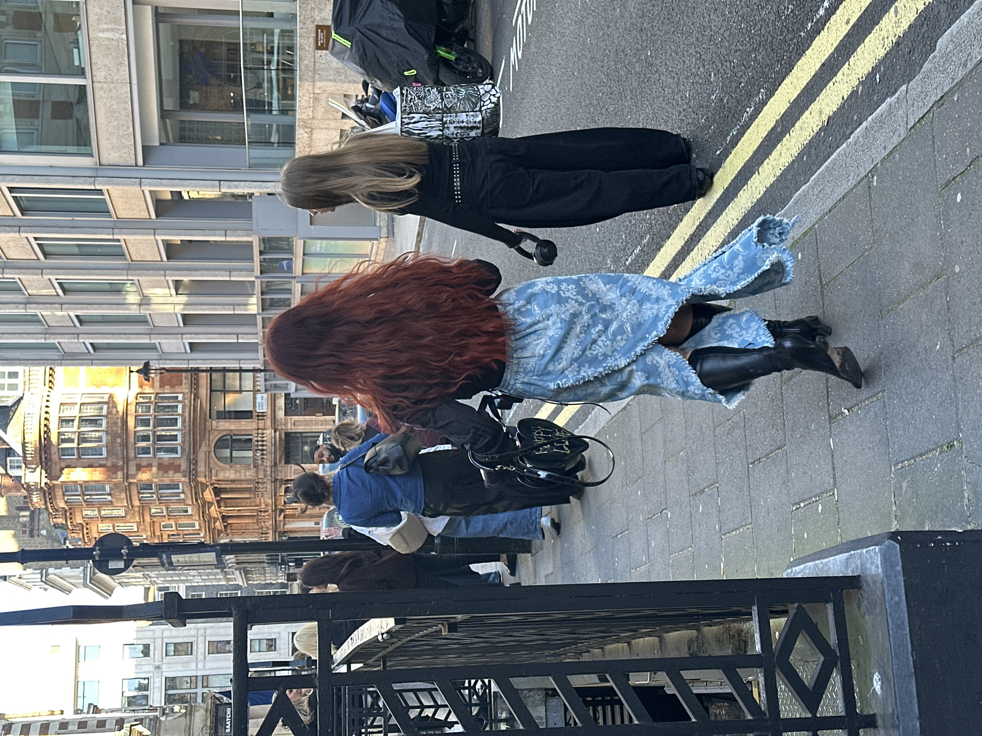This street scene, set in London, United Kingdom, captures pedestrians walking on a pavement alongside a road. In the foreground, viewed from behind, are two women. The woman on the right has long, vibrant red hair and is wearing a light blue, frayed-hem skirt or dress with a white patterned design, paired with black long boots. She carries a black heart-shaped handbag with partially visible white text. To her left, another woman with long blonde hair is dressed in a black long-sleeved top, black trousers, and a studded belt, holding black headphones in her right hand.
Several other individuals are visible walking further down the street. The road surface features prominent double yellow lines, common in the UK. On the far left, a modern building with multiple glass windows and light-colored facades rises. In contrast, the right side and background showcase older, multi-story brick and stone buildings with traditional window structures. A black metal railing with diamond-shaped patterns partially frames the bottom left of the image. A motorcycle covered by a black tarp, with a patterned bag (possibly "MOTOWN" visible on it) strapped to it, is parked by the curb. The lighting suggests daytime, likely with diffused sunlight or an overcast sky. No particular emotions are discernible from the subjects. A distant sign has partially visible text, possibly reading "SAATCHI".