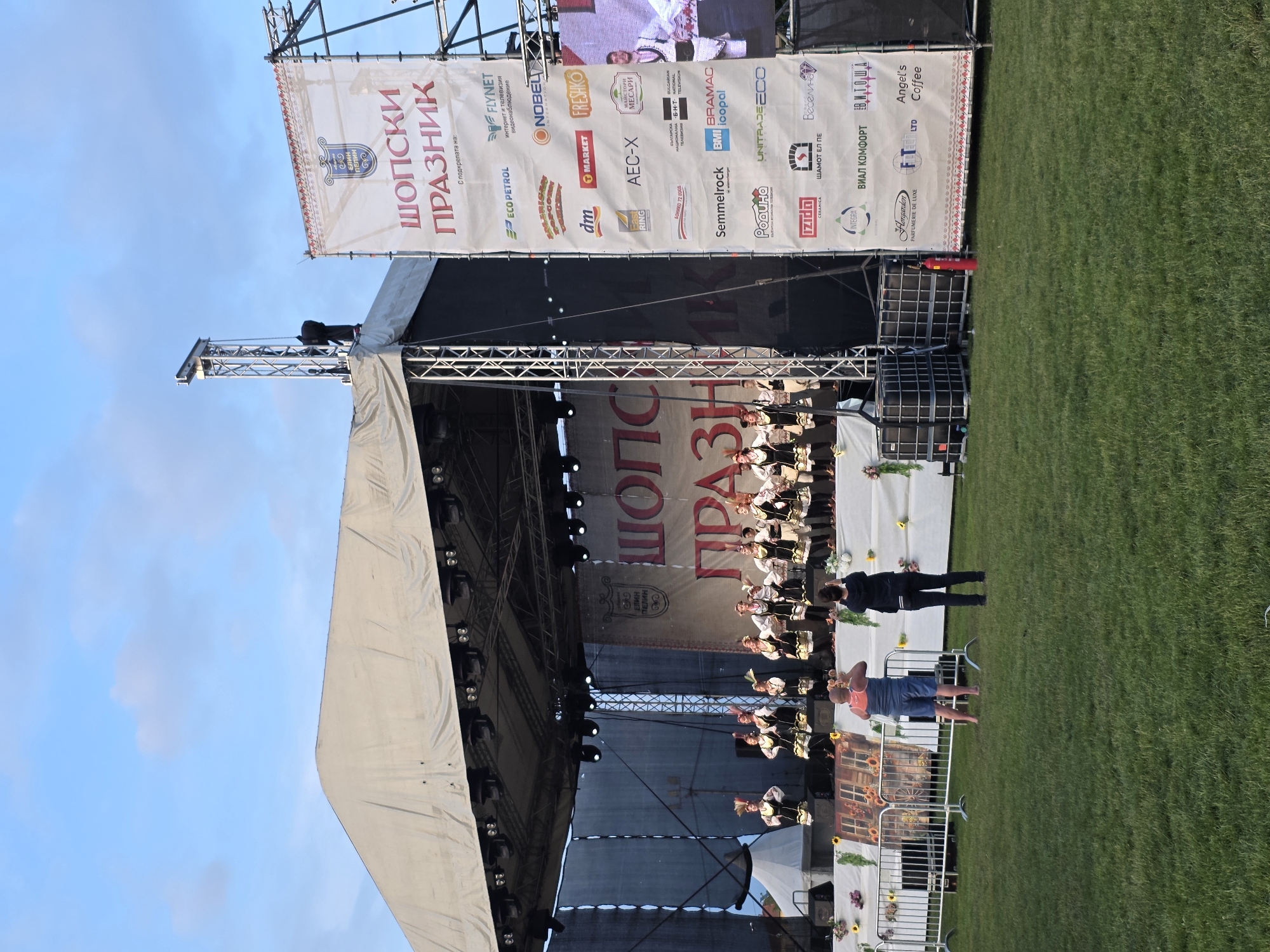 An outdoor cultural festival, identified as the "Shopluk Festival" in Elin Pelin, Bulgaria, is captured during daylight hours under a clear blue sky with scattered white clouds. A large temporary stage is set up on a grassy field. The stage features a light-colored tarp roof, a robust metal truss system supporting numerous stage lights, and a dark backdrop. Approximately a dozen performers, dressed in elaborate traditional Bulgarian folk costumes featuring white blouses, black vests, sashes, and distinctive headwear, are visible on stage, appearing to be in mid-performance. The stage is decorated with floral arrangements, including sunflowers, and a decorative panel depicting a rustic scene.
To the right of the stage, a tall vertical banner framed by metal scaffolding displays the event title "ШОПСКИ ПРАЗНИК" (Shopluk Festival) in red Cyrillic letters, along with a crest featuring "ЕЛИН ПЕЛИН" (Elin Pelin). Below the title, the banner lists "С подкрепата на:" (With the support of:) followed by a dense array of corporate and institutional sponsor logos, including ECO PETROL, FLYNET, NOBEL INTERNATIONAL, dm, MARKET, FRESHKO, Bulgarian National Television (BNT), Semmelrock, BMI icopal, and others. Partially visible behind this banner is a large screen displaying a close-up of a performer.
In the foreground, a neatly mown green grass field extends from the viewer to a metal crowd barrier. Two individuals stand in front of the barrier, observing the performance. The person on the left is a barefoot male wearing a blue and orange sleeveless top and blue shorts, holding a phone or camera to film the stage. The person on the right is a female wearing a black long-sleeved top and black pants, also holding a phone or camera, pointed towards the stage. Behind the barrier, to the left, two black Intermediate Bulk Containers (IBC tanks) and a red fire extinguisher are visible.