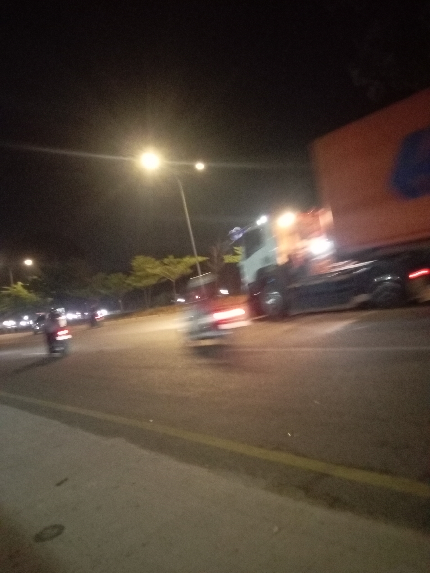 A night scene shows a busy street in Batam, Indonesia, with several vehicles and pedestrians. The street is illuminated by streetlights, creating a somewhat hazy atmosphere. In the foreground, the pavement and a yellow line marking a lane are visible. Several motorcycles are in motion, their lights blurred by the low-light conditions and movement. Further down the road, a large truck with an orange cargo container is prominent, its headlights and taillights also contributing to the illumination. Trees line the side of the road, their foliage illuminated by the artificial lights. The overall impression is one of urban activity at night.