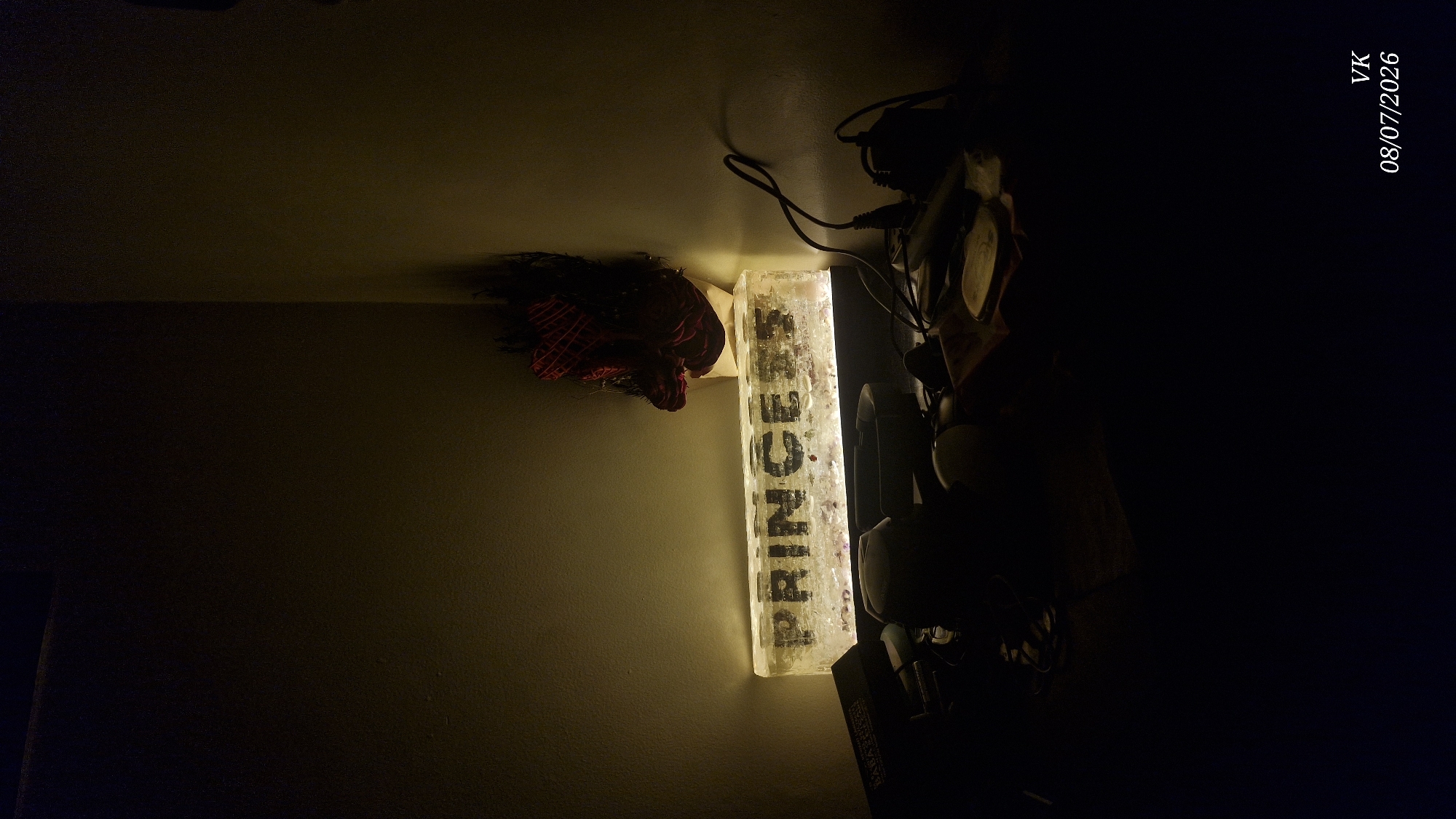 The image depicts a dimly lit corner of a room, possibly a shelf or table, in Vrindavan, India. A prominent rectangular object, seemingly made of frosted glass or resin, is illuminated from within, displaying the word "PRINCESS" in black, block letters. The text appears slightly distressed and embedded within the material. To the right of this illuminated object, a decorative item with red and black feathery elements is placed, resting on what appears to be a mannequin head.
In the foreground, the scene is largely obscured by shadows. Various objects are scattered on a surface, including what look like electronic devices, cables, and other indistinct items. One object in the lower left corner appears to be a dark-colored box or book. The overall lighting is low, casting long shadows and highlighting the illuminated "PRINCESS" sign. The wall in the background is a light, textured surface. A watermark in the bottom right corner reads "VK 08/07/2026".
