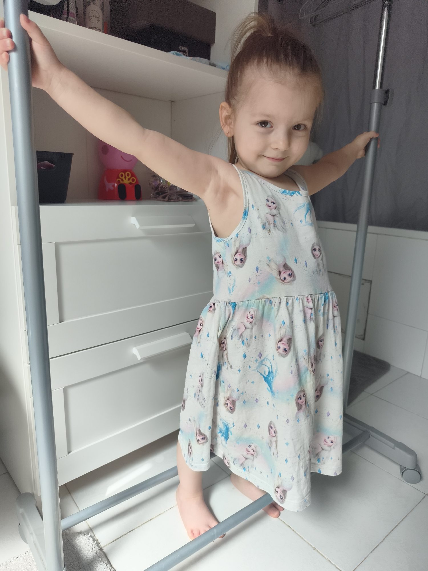 A young Caucasian girl with light skin and brown hair pulled into a high ponytail stands with her arms outstretched, holding onto a gray metal rack. She is wearing a sleeveless white dress with a repeating pattern of cartoon princesses, possibly from the movie Frozen. The girl is barefoot and her feet are positioned on the bottom bar of the rack. She is smiling and looking towards the camera.
The background consists of white furniture with drawers and shelves. On one of the shelves, a pink toy pig is visible, along with other small items. The floor is tiled in white. The environment appears to be indoors, likely a bedroom or closet area. The image was taken in Lagny-sur-Marne, France.