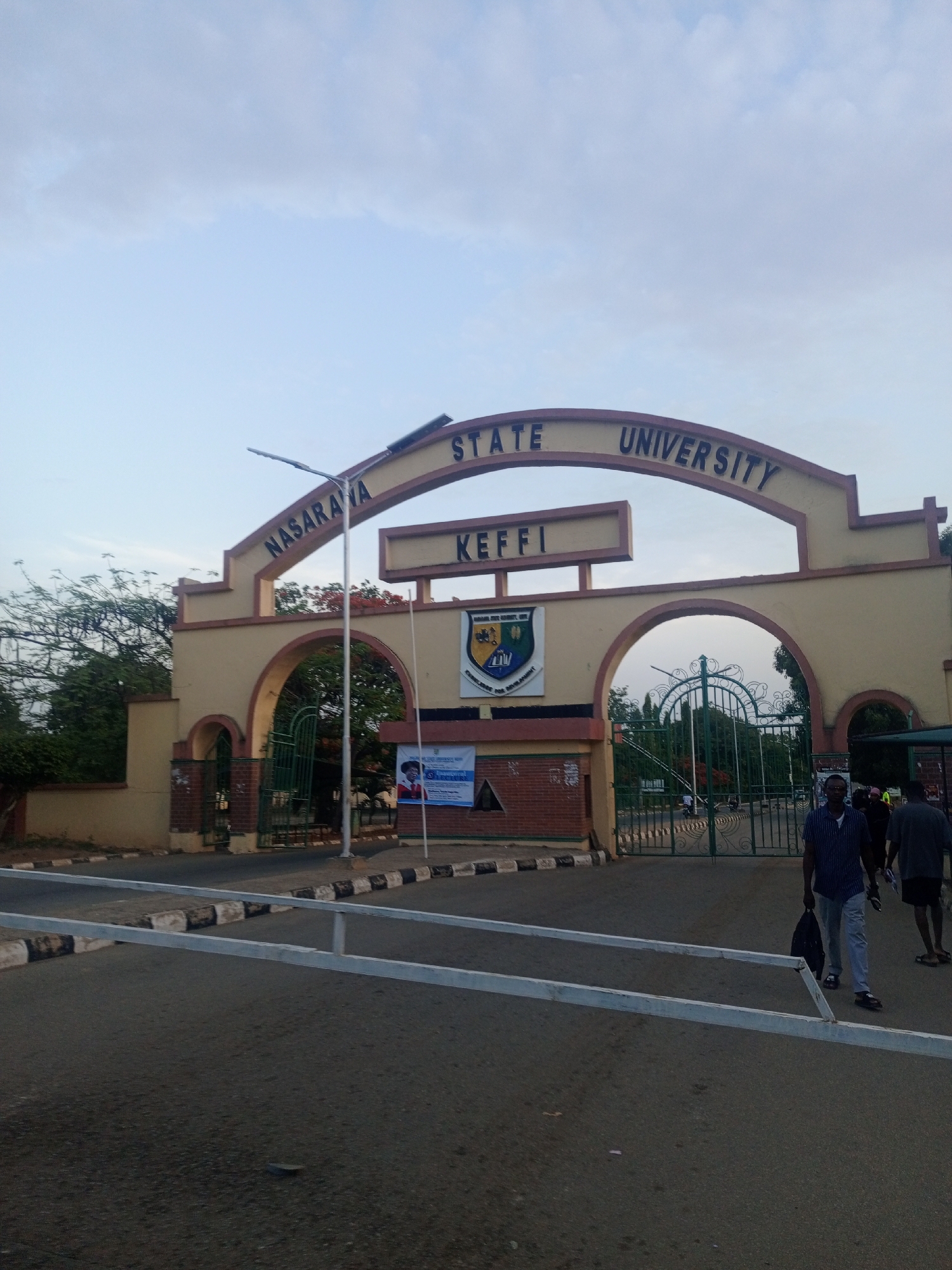 The image displays the main entrance gate of Nasarawa State University in Keffi, Nigeria. The prominent archway structure, painted in a light tan or beige color with darker trim, spans across a roadway. It features three arched openings, with the largest central arch flanked by two smaller ones, behind which green, ornate metal gates are visible, suggesting controlled access.
Above the central arch, the name "NASARAVA STATE UNIVERSITY" is inscribed in dark letters along the curve. Below this, on a horizontal beam, the word "KEFFI" clearly identifies the university's location. A university crest or logo, containing phrases like "UNIVERSITY CHALLENGE FOR DEVELOPMENT" and "UNITY AND SERVICE," is affixed to the central pillar of the gate.
In the foreground, a paved road leads up to the gate, bordered by a curb with distinctive black and white stripes. A white metal barrier, resembling a boom gate arm, is lowered across the road, indicating a restriction on vehicular entry or exit.
Several people are visible near the entrance. In the right foreground, a man in a striped blue shirt and light-colored trousers walks towards the viewer, carrying a dark bag. Behind him, another person is partially seen, and further to the right, a person in a dark t-shirt and shorts walks away. Other individuals can be seen moving within the university grounds behind the gate.
To the left of the central arch, a tall street light pole with two lights stands. Lush green trees and foliage are visible behind and to the left of the gate. On the right side of the gate, a banner or poster with an image of a person and some text is displayed. The sky overhead is bright but overcast with light clouds, suggesting either early morning or late afternoon, characterized by soft, diffused natural light. The scene depicts a typical daily flow of individuals at a university entrance.