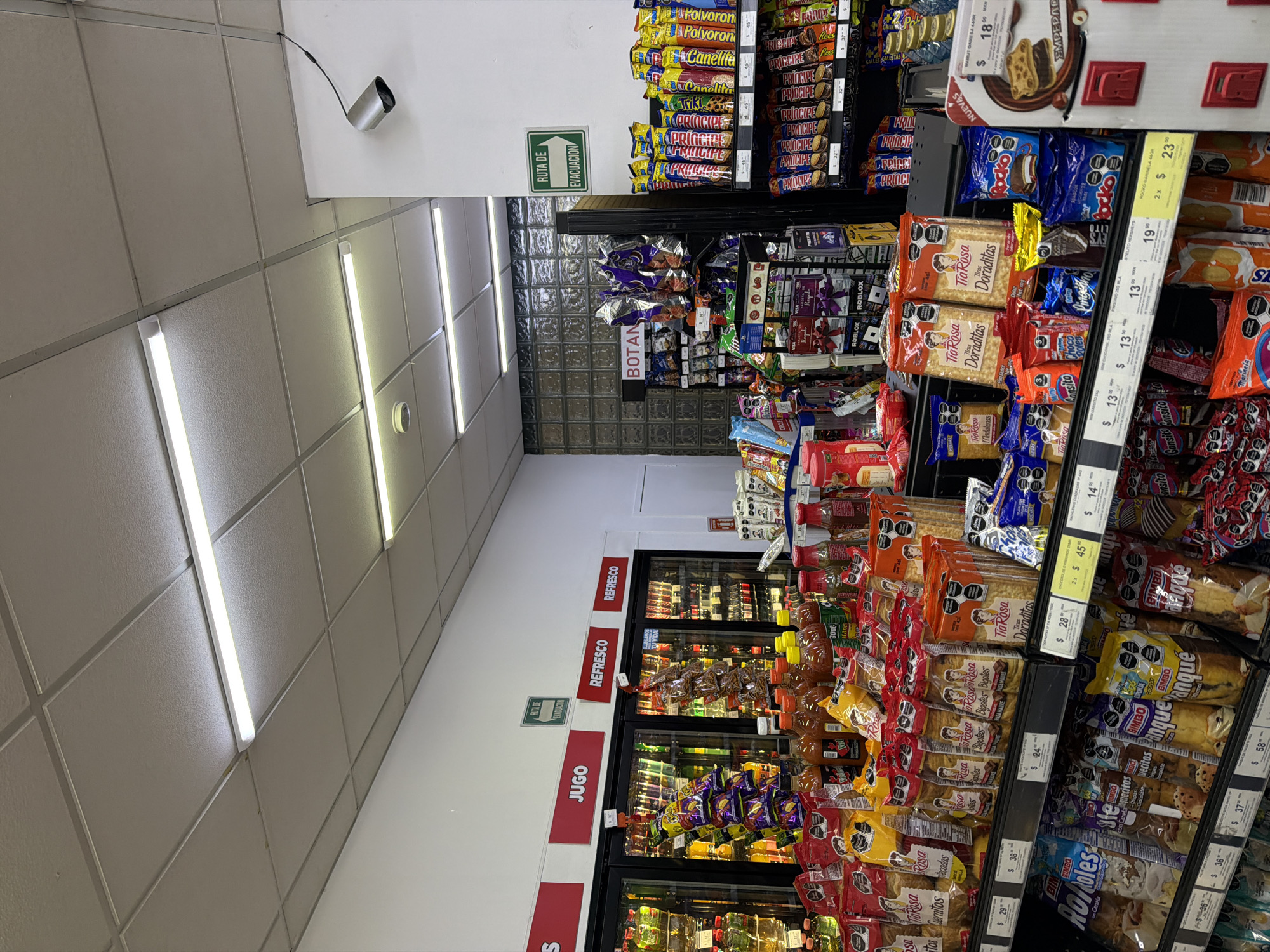 The image depicts the interior of a convenience store in San Andrés Cholula, Mexico. Aisles are stocked with various snacks, candies, and baked goods, including Tía Rosa brand products, cookies, and "Gansito" snack cakes. Refrigerated coolers offer juice and soft drinks. Signs in Spanish indicate categories such as "JUGO" (juice), "REFRESCO" (soda), and "BOTAN" (snacks). A green "RUTA DE EVACUACION" (evacuation route) sign is visible on the wall near the ceiling, alongside a security camera. Pricing and promotional labels are displayed on the shelves. The fluorescent lighting is bright, and the ceiling is tiled.