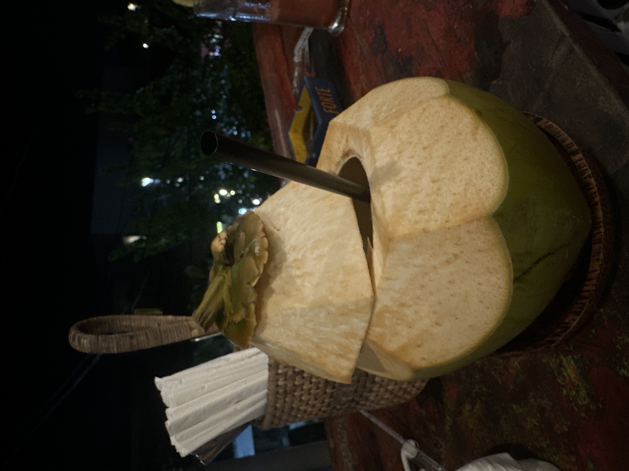 A fresh coconut drink with a metal straw sits on a textured brown surface, partially atop a woven basket holding white napkins. The coconut has been cut to allow access to the liquid inside. To the left, a dark, out-of-focus background suggests an outdoor setting with lights and foliage. A blue and yellow object is visible near the coconut. The scene is likely in North Kuta, Indonesia.