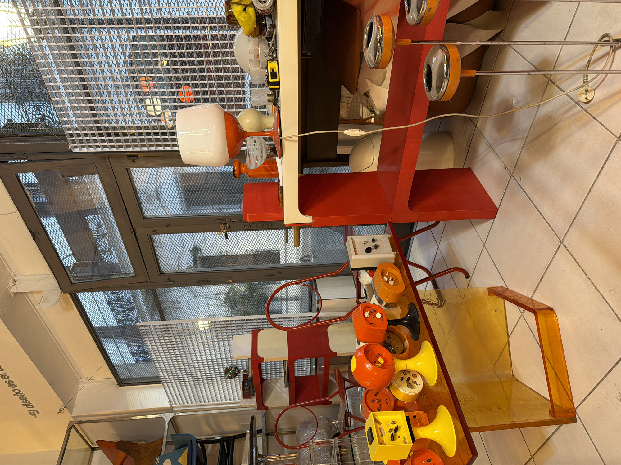 The image captures an interior space in what appears to be a shop or showroom in Buenos Aires, Argentina, as indicated by the phrase "El diseño es el..." partially visible on the wall. The focus is on a collection of vintage or retro design objects, primarily lighting fixtures and furniture. There is an orange and white table lamp with a square shade, vintage orange and chrome sconce lamps, and a table with several disassembled lamps in yellow, orange, and black on it. A red shelving unit displays additional white objects. The furniture includes red chairs with curved backs and an orange acrylic chair with a transparent seat and armrests. The interior has a grid-patterned window covering. The scene shows objects arranged for display or sale.