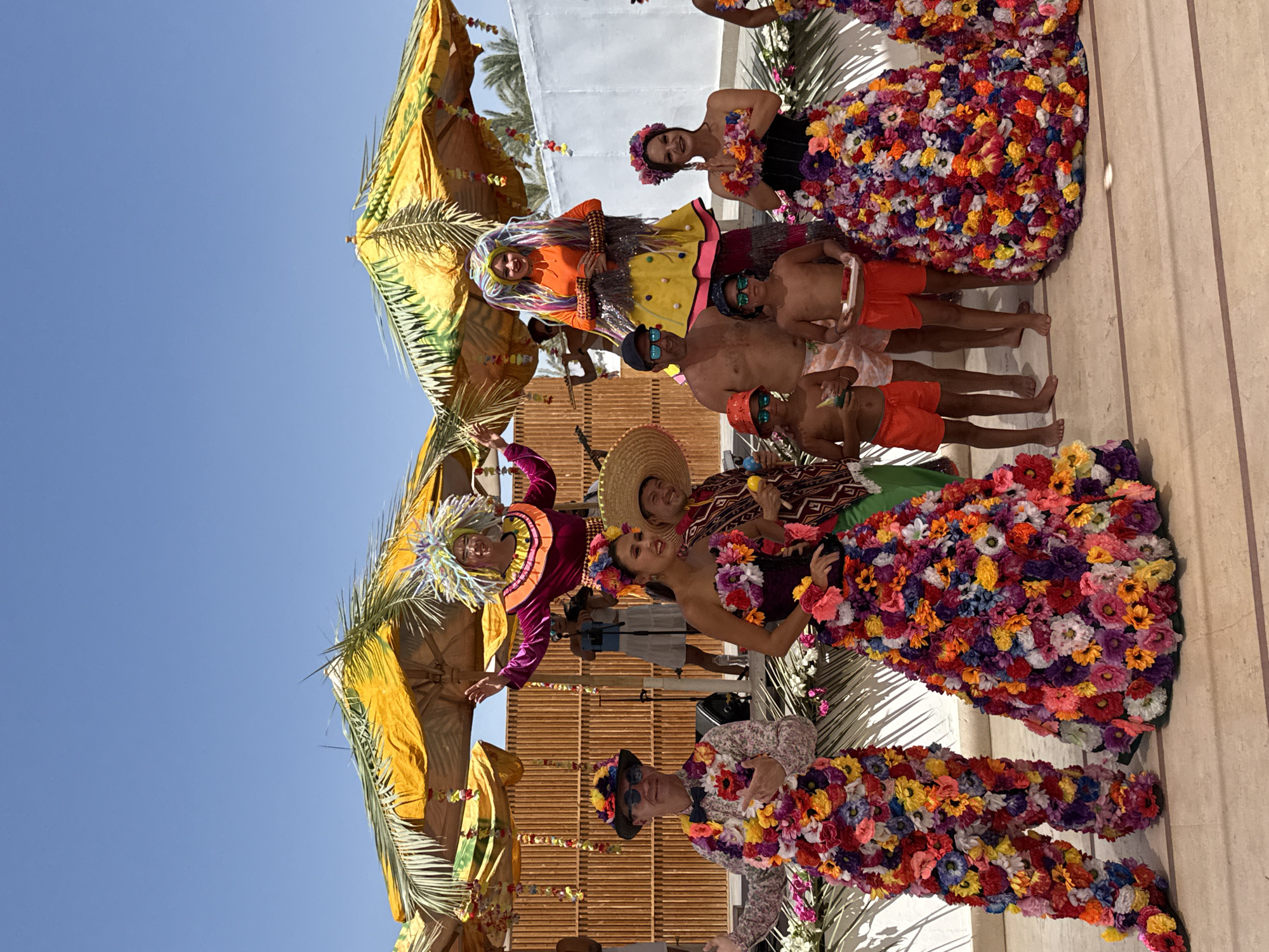 In Sharm El Sheikh, Egypt, a group of people are posing for a photo outdoors under a clear blue sky. Two individuals, one dressed as a stilt walker with a large, colorful wig and a bright pink and yellow outfit, and another in a similar stilt costume with a colorful wig and an orange top with a yellow skirt, stand prominently. Several other individuals are dressed in vibrant, flower-covered costumes, with some wearing hats and sunglasses. Two young boys in swim trunks are also present. They are standing on a tiled surface, with thatched umbrellas and palm fronds providing shade. The backdrop includes a wooden fence and a white wall. The scene appears to be a festive event or performance.
