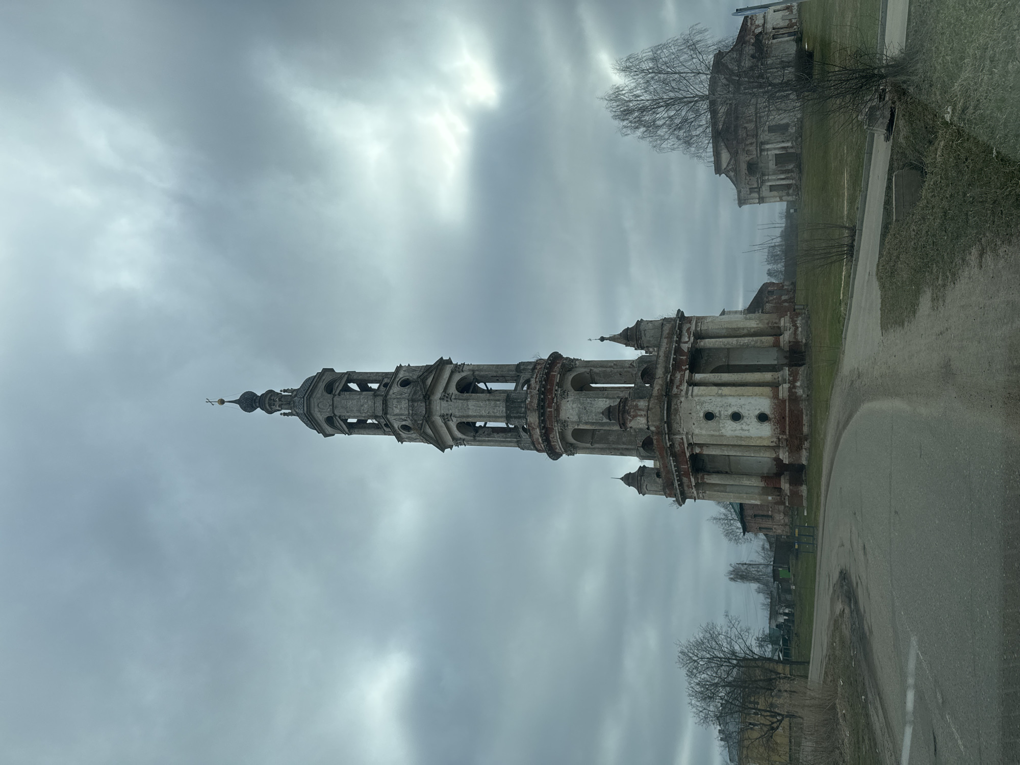 The image depicts the ruins of St. Nicholas Church in Kladovitsy, Russia, under a cloudy sky. The church's bell tower is the most prominent feature, appearing dilapidated and incomplete, with multiple tiers and arched openings. A ruined building stands in the background to the right. A road runs along the right foreground, and trees are visible on the periphery. The general environment suggests a state of decay and neglect.
