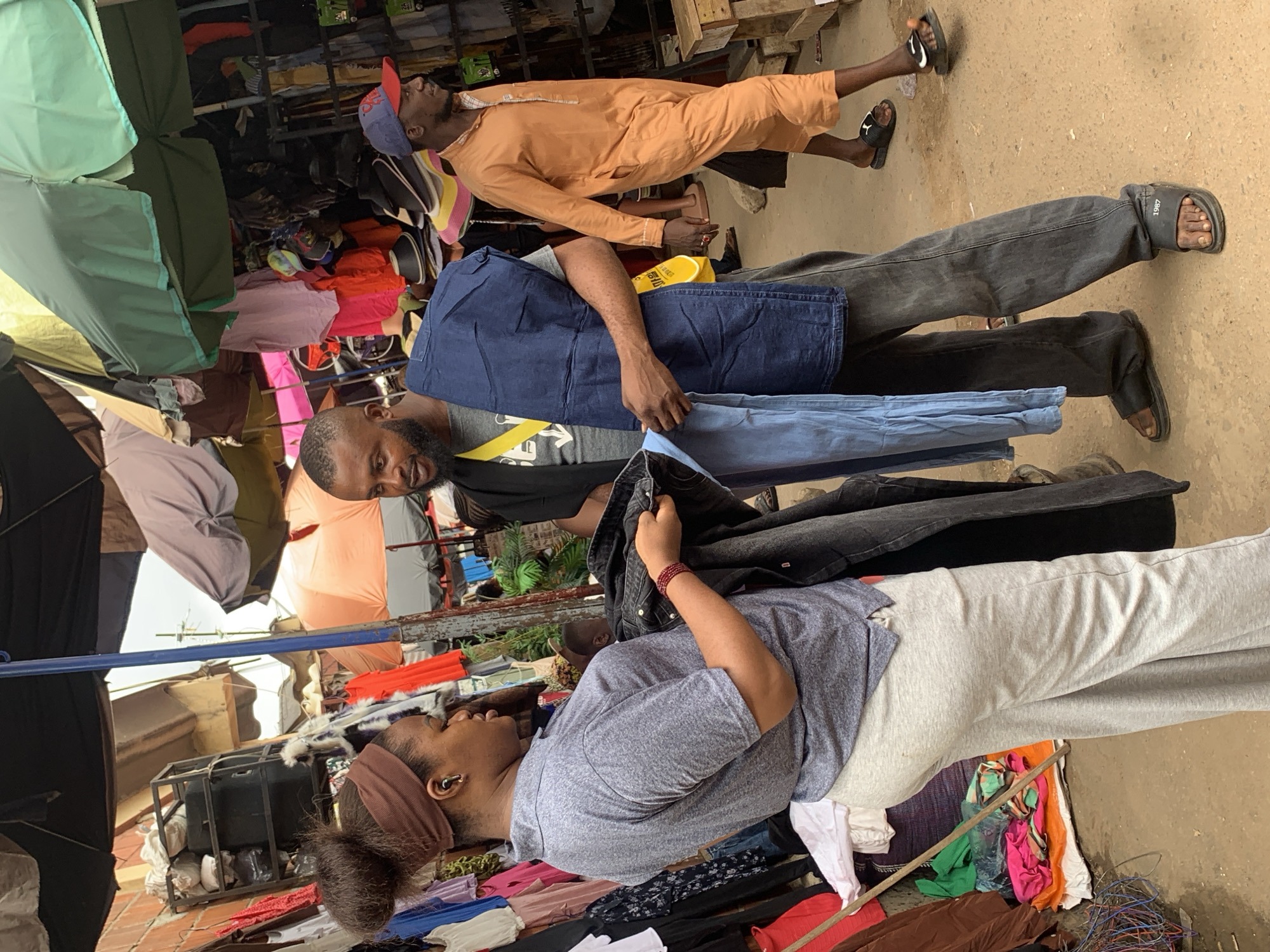The image depicts a street market scene, possibly in Abuja, Nigeria. In the foreground, a woman with her hair in a bun and wearing a brown headband and a grey polo shirt is inspecting dark colored clothing, possibly denim jeans. She is wearing a red beaded bracelet on her left wrist. To her right, a man with a beard, wearing a grey t-shirt and a dark vest with a yellow strap, is holding up folded blue denim fabric, likely a pair of jeans, towards her. He is wearing dark grey pants and black slides with the number "1987" visible on the strap. Further to the right, a man in an orange traditional outfit is walking away from the viewer, wearing black slides. The background shows stalls with various goods, including clothes and hats, under colorful umbrellas. The ground is dusty and appears to be unpaved. The scene suggests a bustling commercial environment.