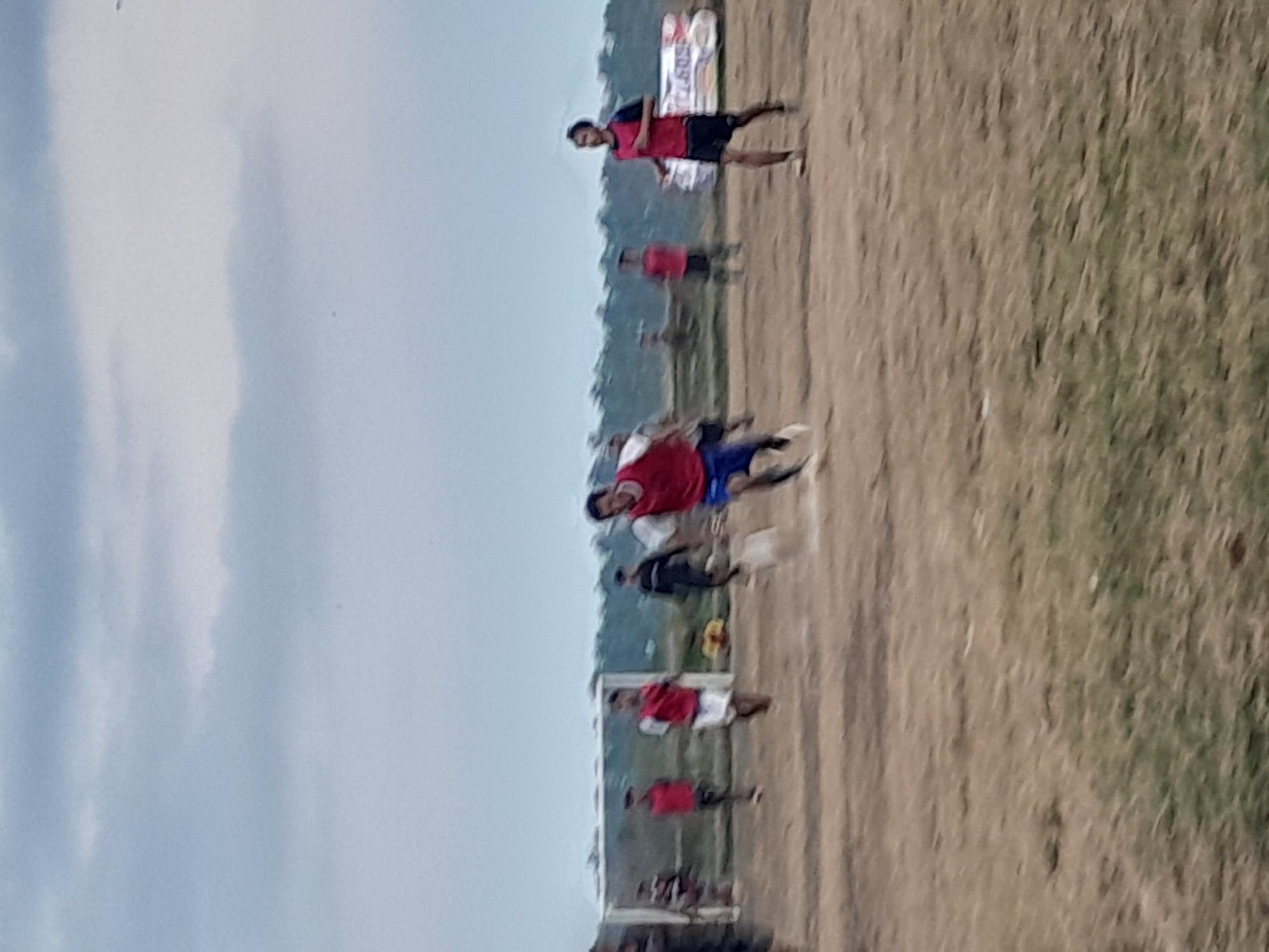 The image depicts a soccer game on a dirt field, likely in Genteng, Indonesia, under an overcast sky. Several players, most wearing red jerseys and dark shorts, are in motion. One player in the foreground, wearing a red vest and blue shorts, is running towards the viewer, appearing to be in the middle of a play. Another player in a red vest and white shorts is also running, his face obscured. In the background, a soccer goal is visible, with other players scattered around the field and a banner with text is present behind some of the players. The ground is dry and dusty with patches of sparse grass. The overall scene suggests an informal or local soccer match.