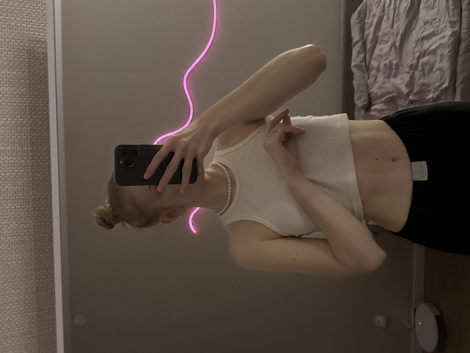 An indoor mirror selfie depicts a young adult female located in a residential space within the city of Perm, Russia. The subject, with blonde hair styled in a top bun, is seen from the mid-chest down, with her face partially obscured by a dark-colored smartphone held in her right hand, which is taking the photograph. She is wearing an off-white ribbed crop top, exposing her midriff, and dark-colored trousers with a light-colored belt buckle visible on her left hip. A single-strand pearl necklace adorns her neck, and a ring is visible on the ring finger of her left hand, which rests near her abdomen. Behind the subject, a wavy pink neon light strip is affixed to a light gray wall. To the lower left of the frame, a white disc-shaped robot vacuum cleaner is positioned on a light brown floor, beneath a white wall outlet. To the right, a bed with wrinkled light purple bedding is partially visible. The ceiling above is textured and light-colored. The scene is illuminated by soft ambient light and the glow of the neon strip.