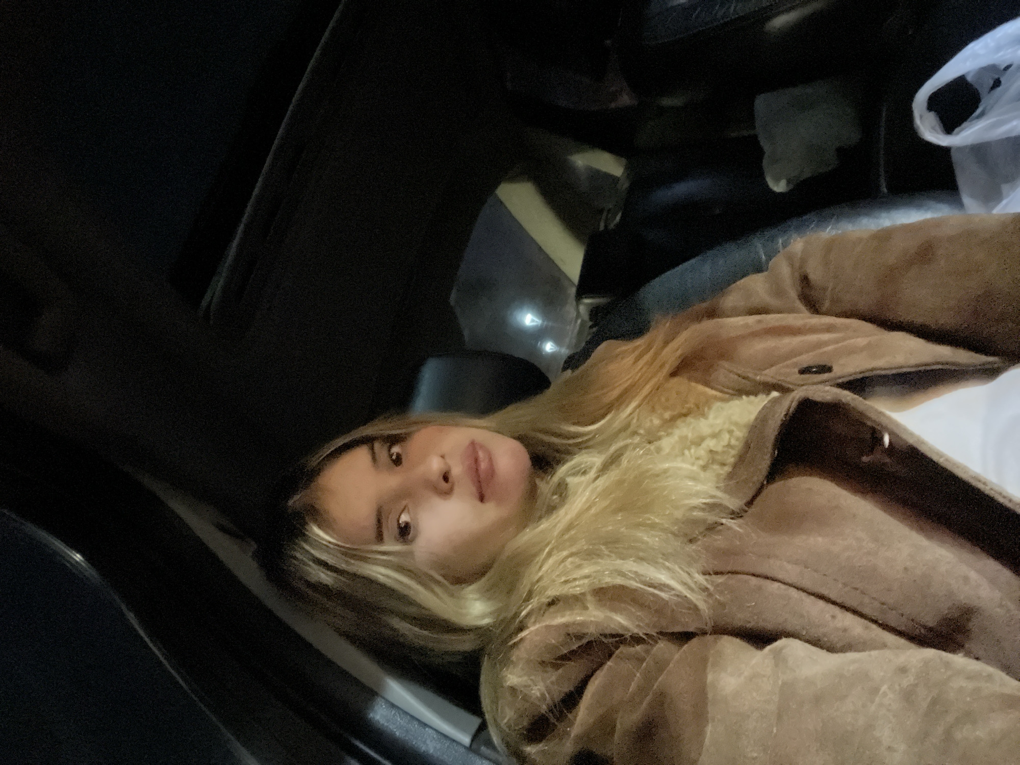 A person with light hair, wearing a brown jacket with a fur collar, is sitting in a vehicle. The interior of the vehicle is dark. The location is not discernible from the image. Therefore, specifying the city of San Fernando del Valle de Catamarca, Argentina is impossible.