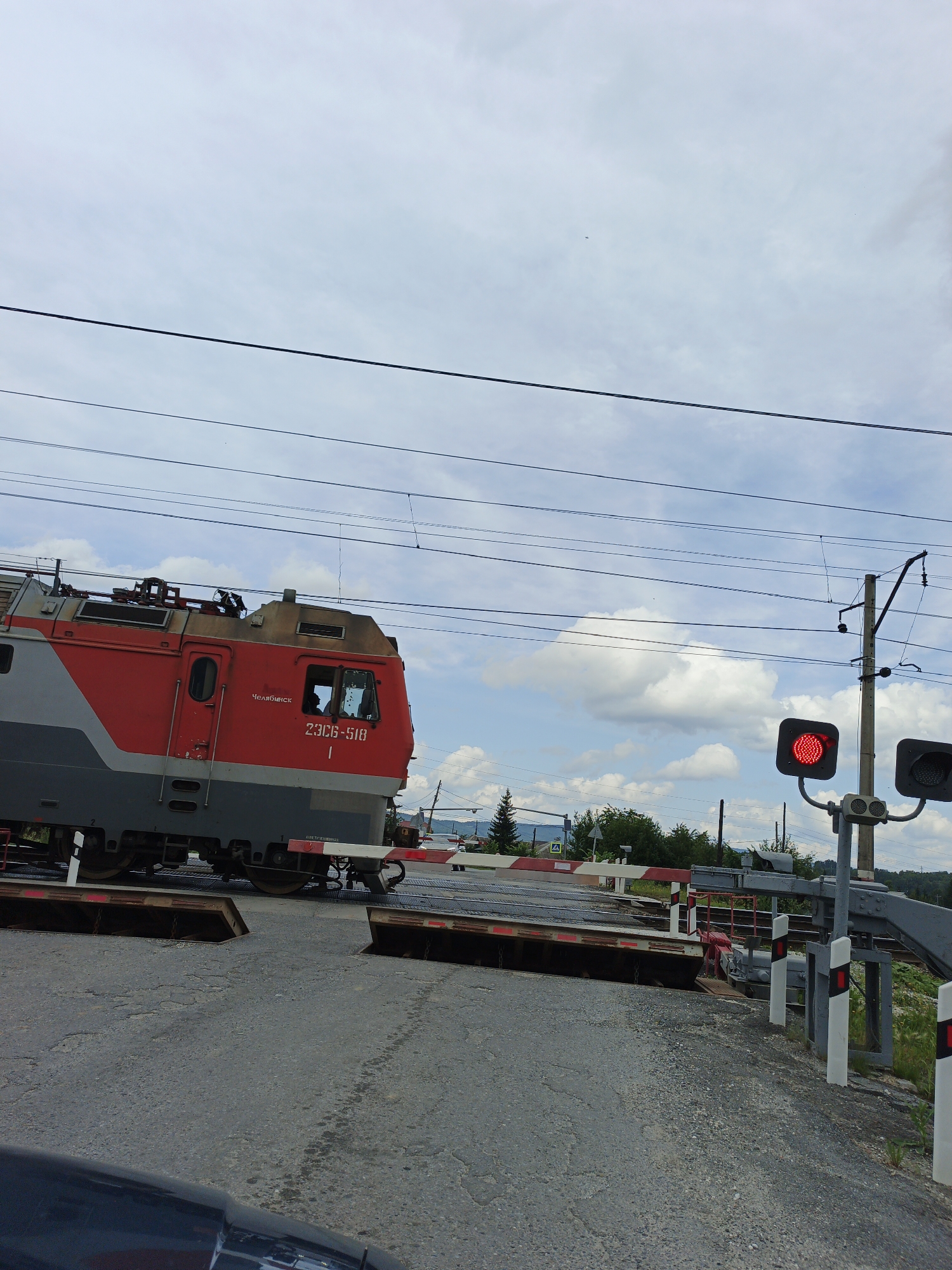 In Medvedyovka, Russia, a red and gray electric train, designated as 23CB-518, is stopped at a railroad crossing. The train's number and "ЧЕЛЯБИНСК" (Chelyabinsk) are visible on its side. The crossing is equipped with red and white barriers, and a red signal light is illuminated, indicating a stop. Power lines stretch across the cloudy sky, with a pole supporting the traffic signals. The ground at the crossing is paved with asphalt and gravel. The surrounding environment includes trees and distant hills. The image appears to have been taken from a vehicle, as the hood of a car is visible in the foreground.